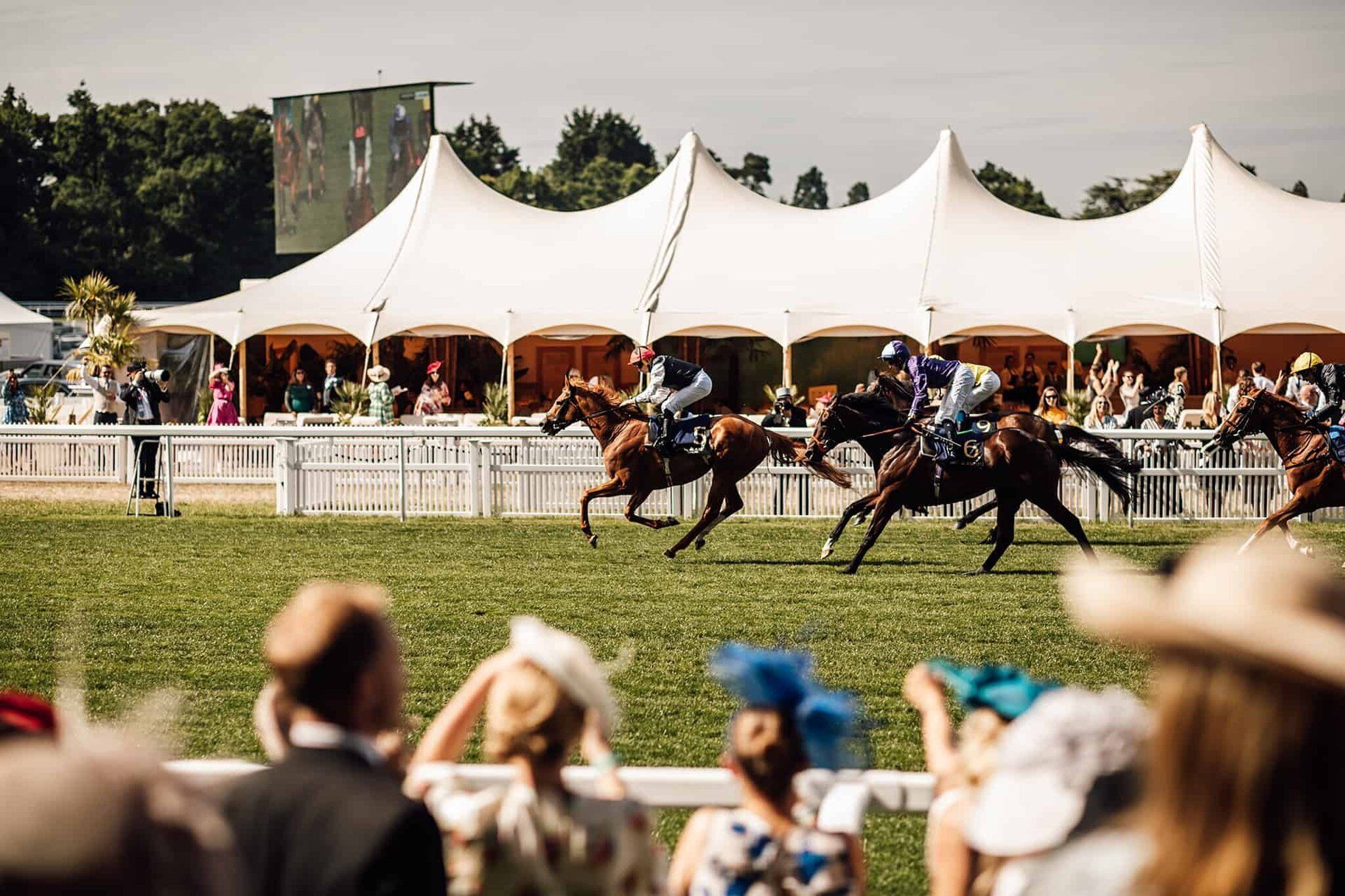 A horse race with jockeys on horses, spectators in the foreground, and tents in the background.
