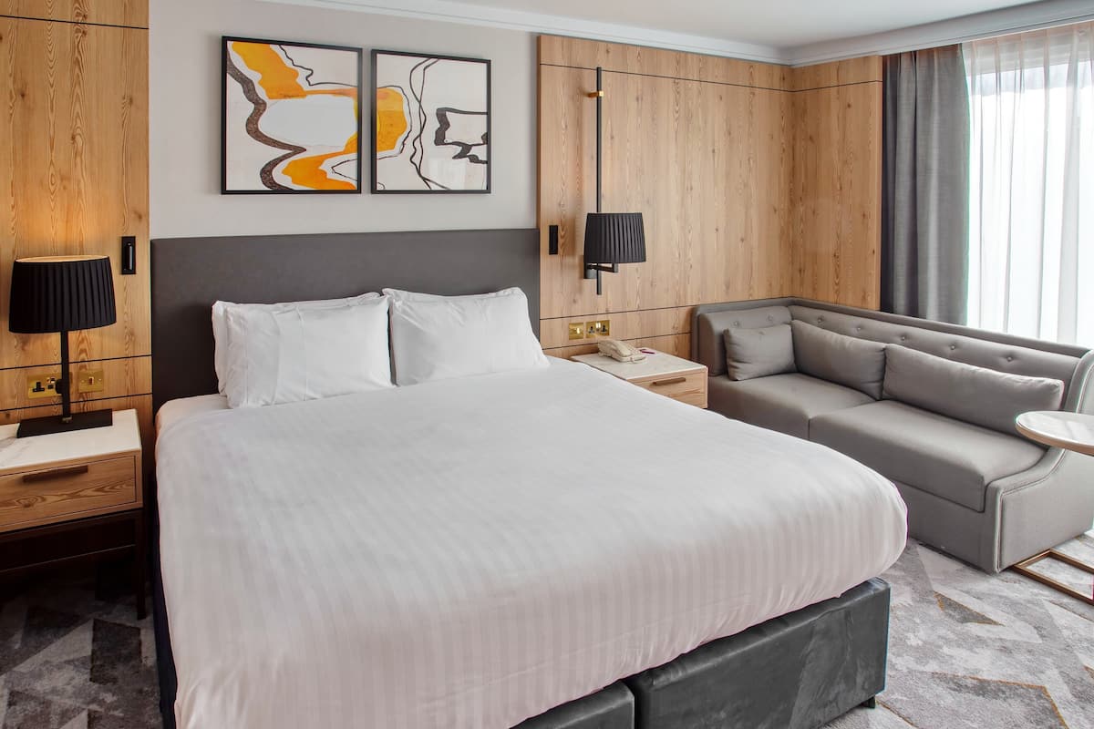 Une chambre d’hôtel avec un grand lit, un canapé, une table de chevet avec une lampe et une lampe fixée au mur. Il y a un tableau au mur et une fenêtre avec des rideaux.