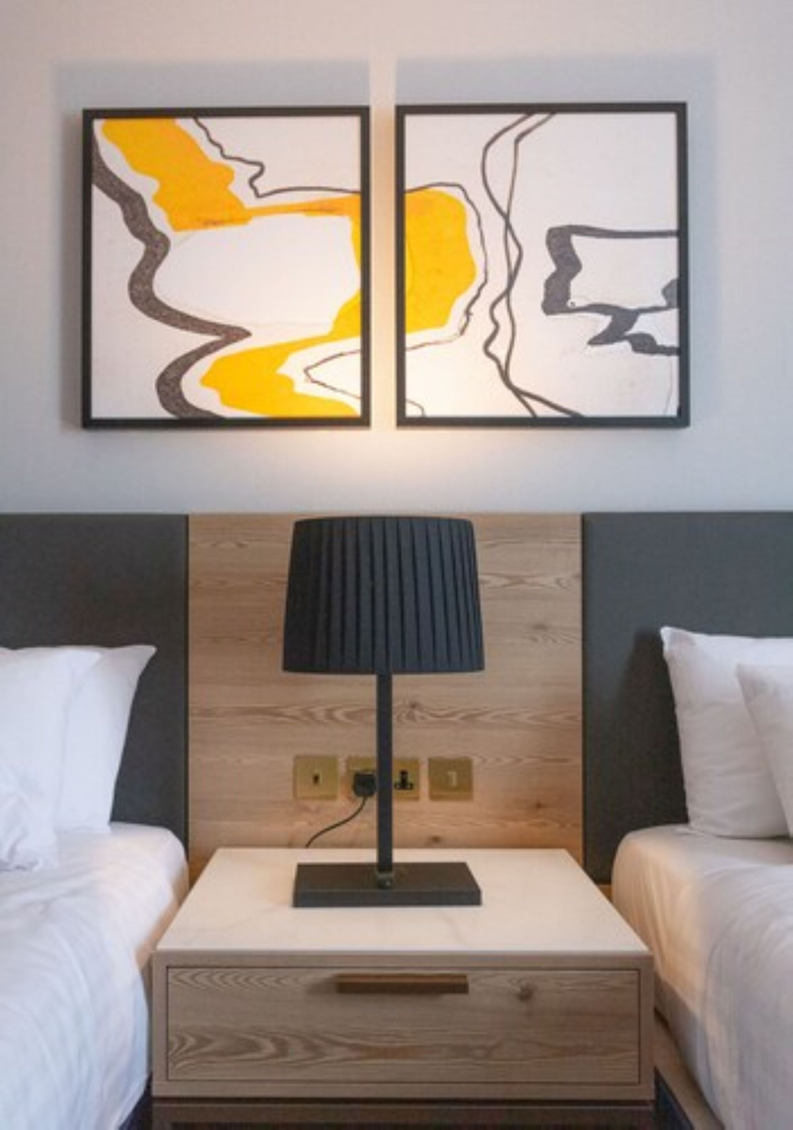 A hotel room with two beds, a wooden headboard, a bedside table with a lamp, and abstract artwork on the wall.