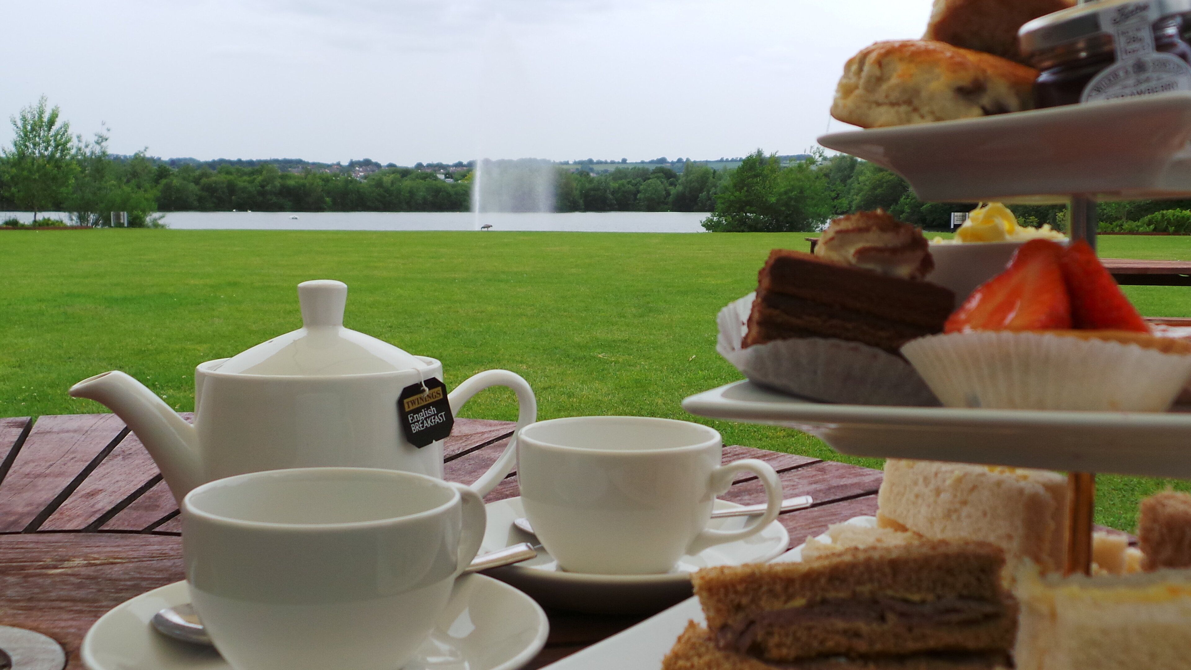 Una disposición para picnic con tetera, tazas y un soporte escalonado para postres sobre una mesa de madera, con vistas a un lago al fondo.