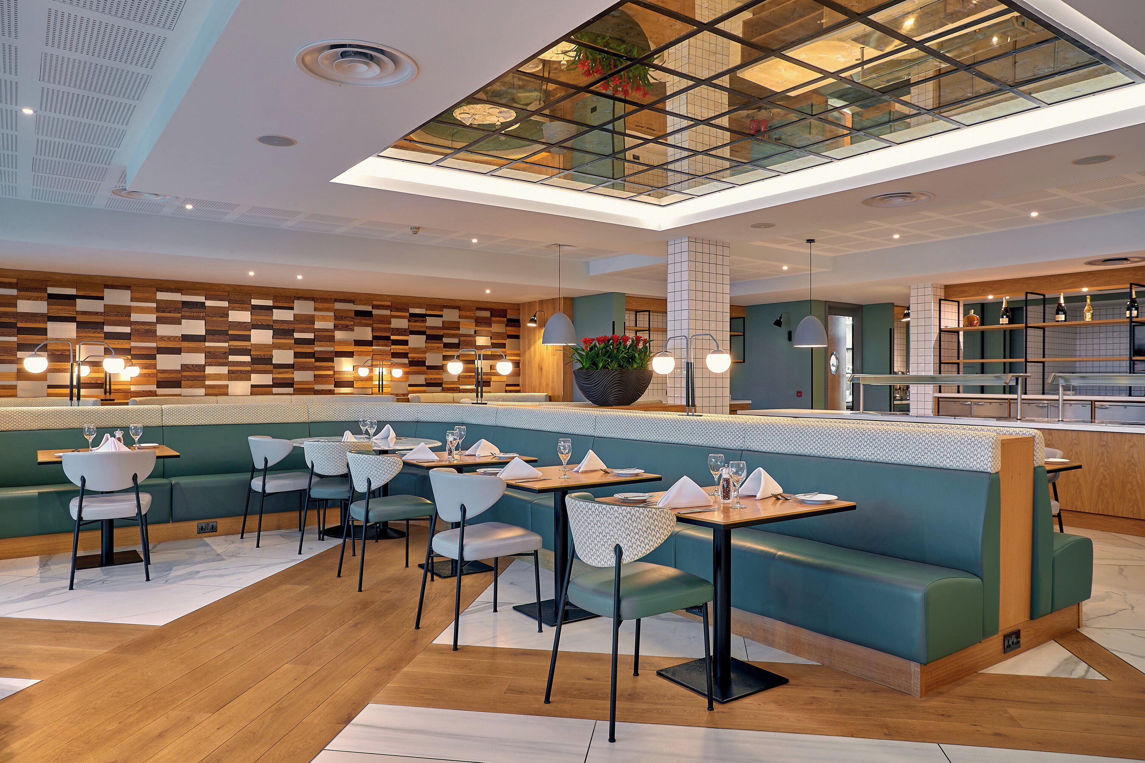 A modern restaurant with a high ceiling, wooden panel wall, and a glass ceiling. Tables and chairs are arranged for dining.