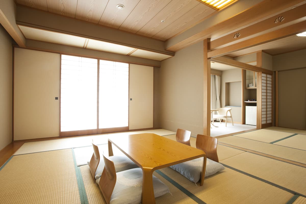 Une pièce traditionnelle japonaise avec un sol recouvert de tatamis, une table en bois et des coussins, de grandes portes coulissantes et une vue sur une autre pièce.