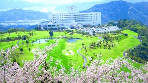 Ein modernes Hotel mit einem "Js"-Schild, umgeben von einem üppigen Golfplatz und Bergen.