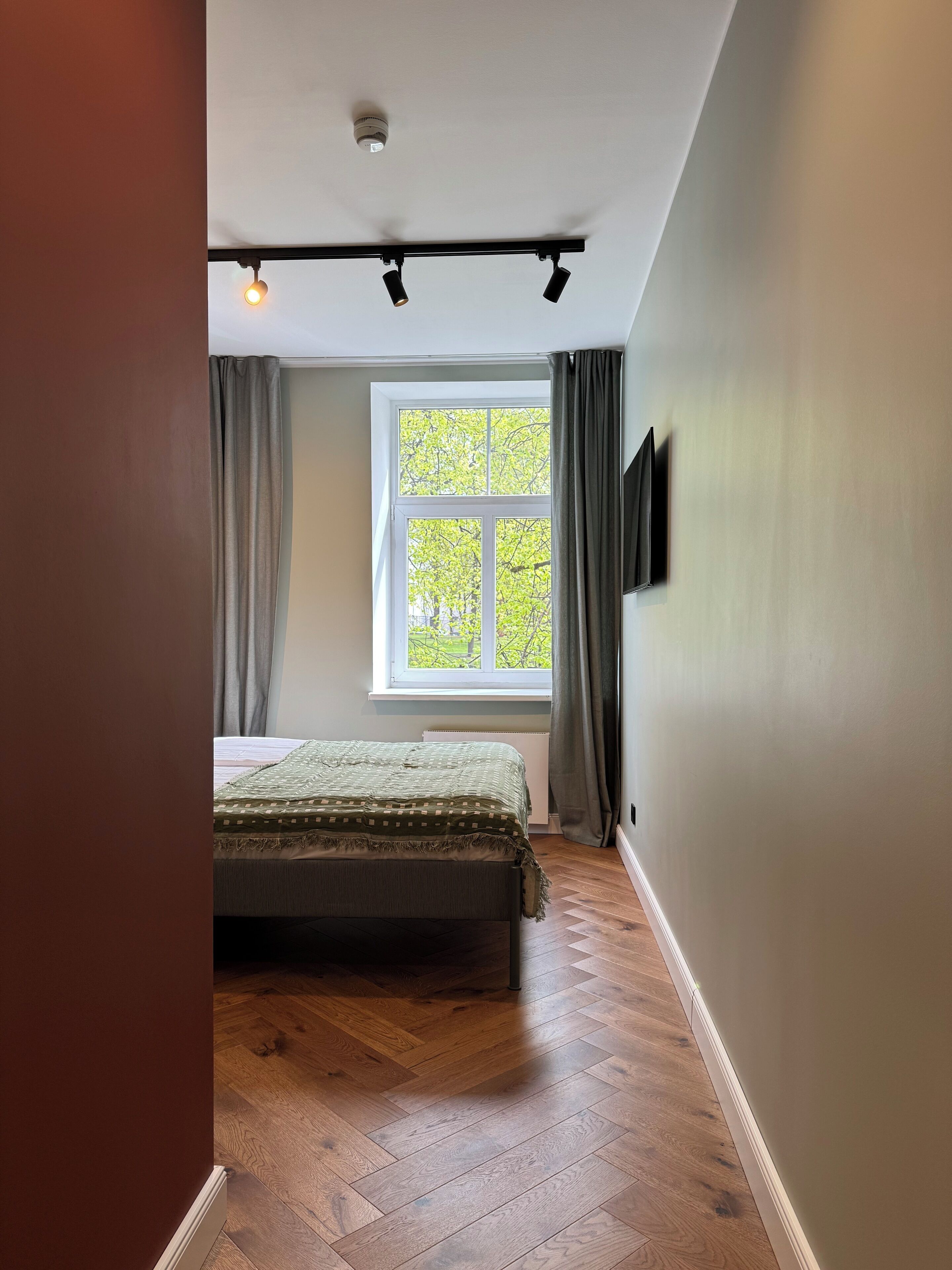 Un dormitorio con cama, ventana con cortinas, una lámpara de techo y televisor montado en la pared.