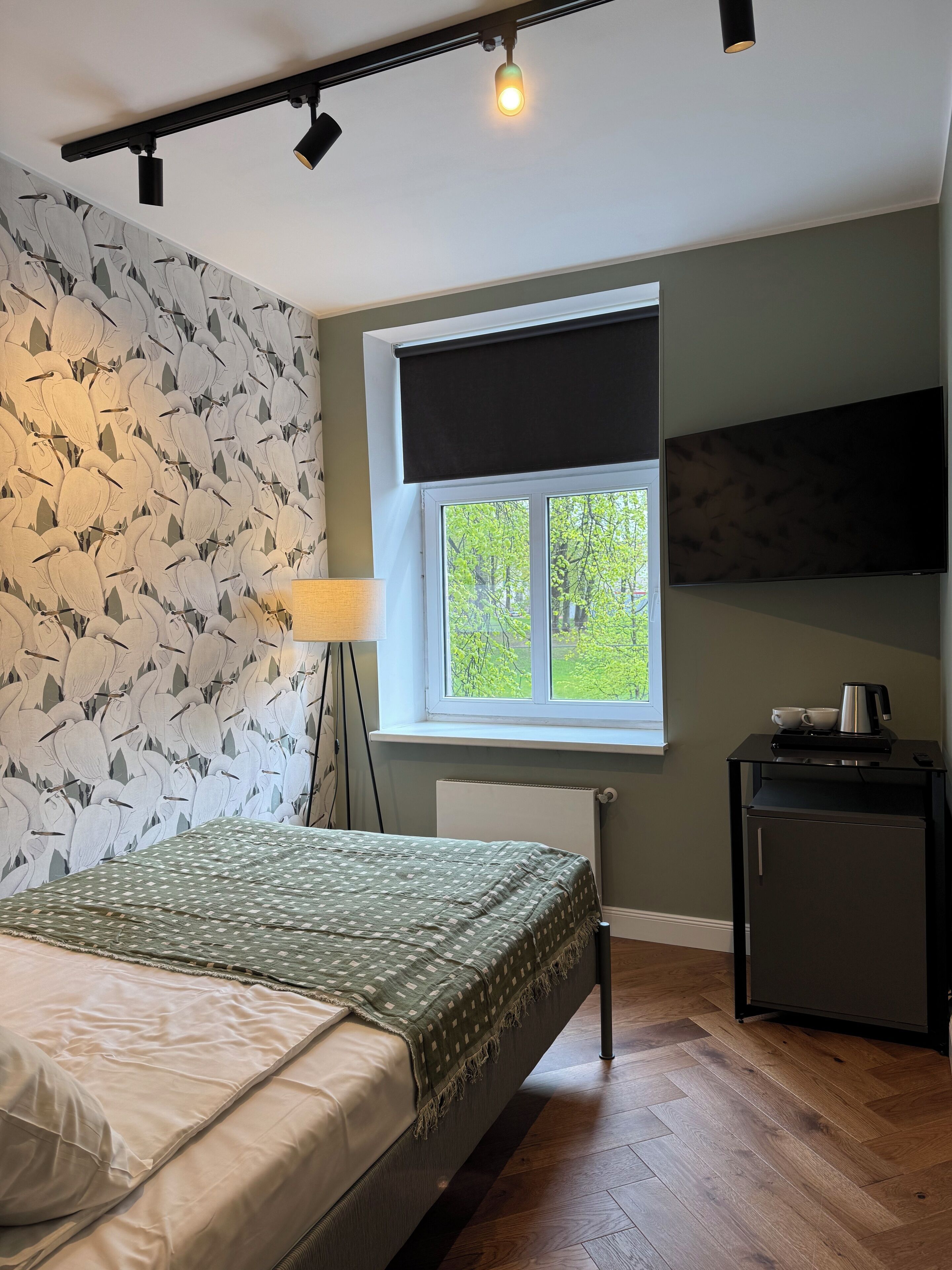 Quarto com cama, janela com vista para vegetação, radiador, escrivaninha com chaleira e TV na parede.