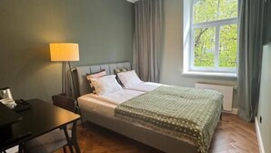 A bedroom with a bed, a desk, a lamp, and a window with curtains. - Hotel Avitar (Riga)