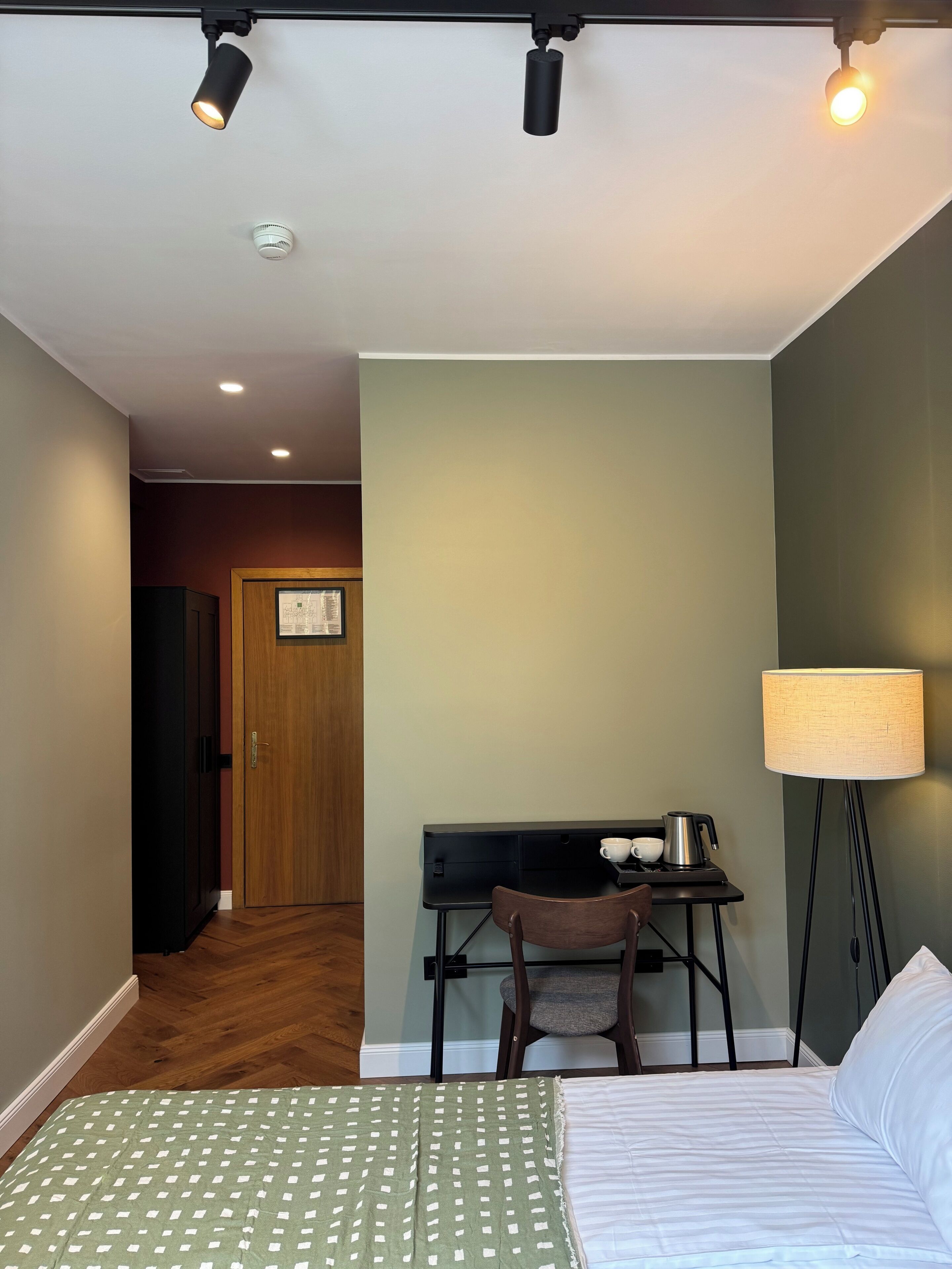A hotel room with a bed, a desk with a chair, a lamp, and a door leading to another room.
