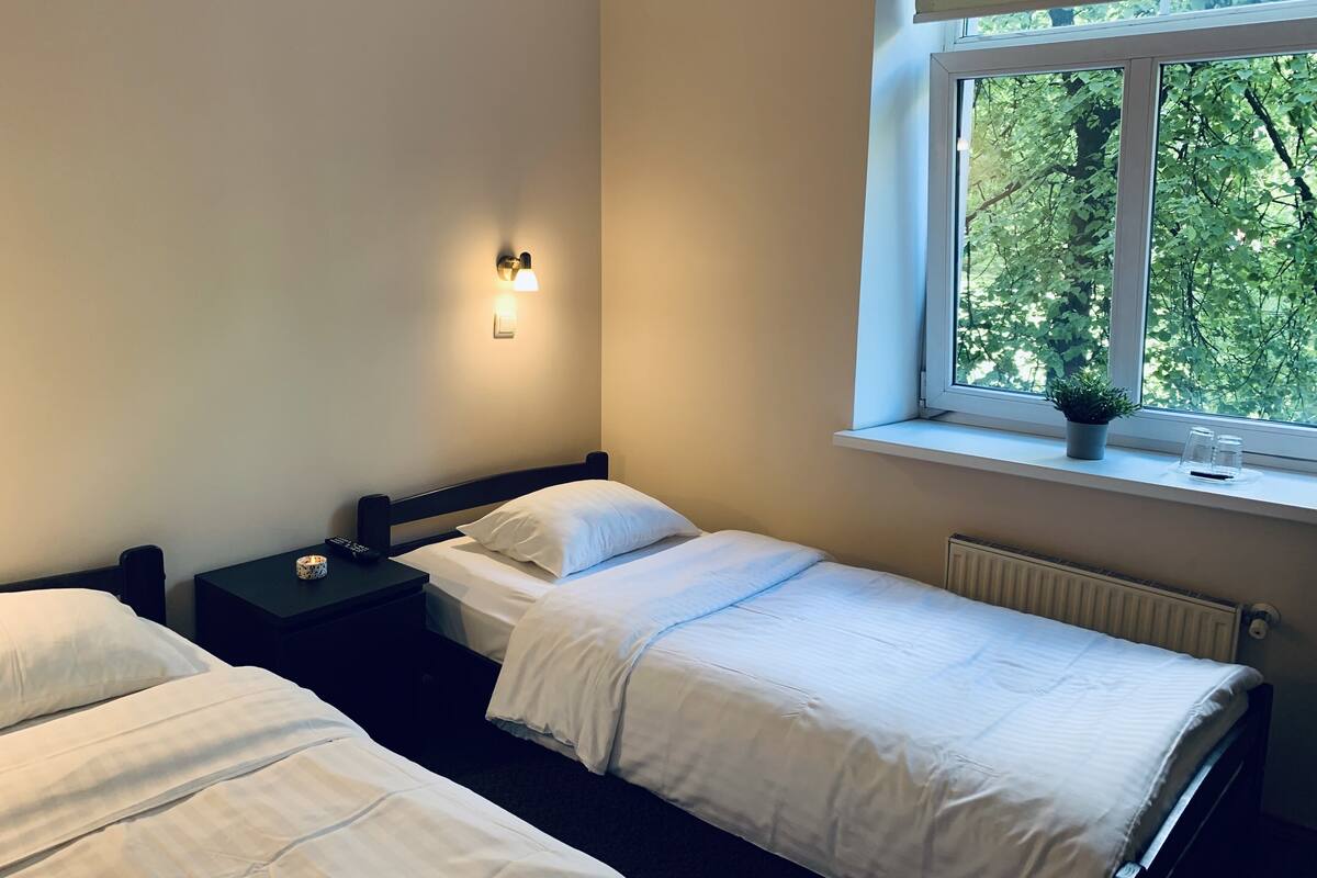 Ein Schlafzimmer mit zwei Einzelbetten, einem Nachttisch mit Lampe, einem Fenster mit Blick auf Bäume und einem Heizkörper.