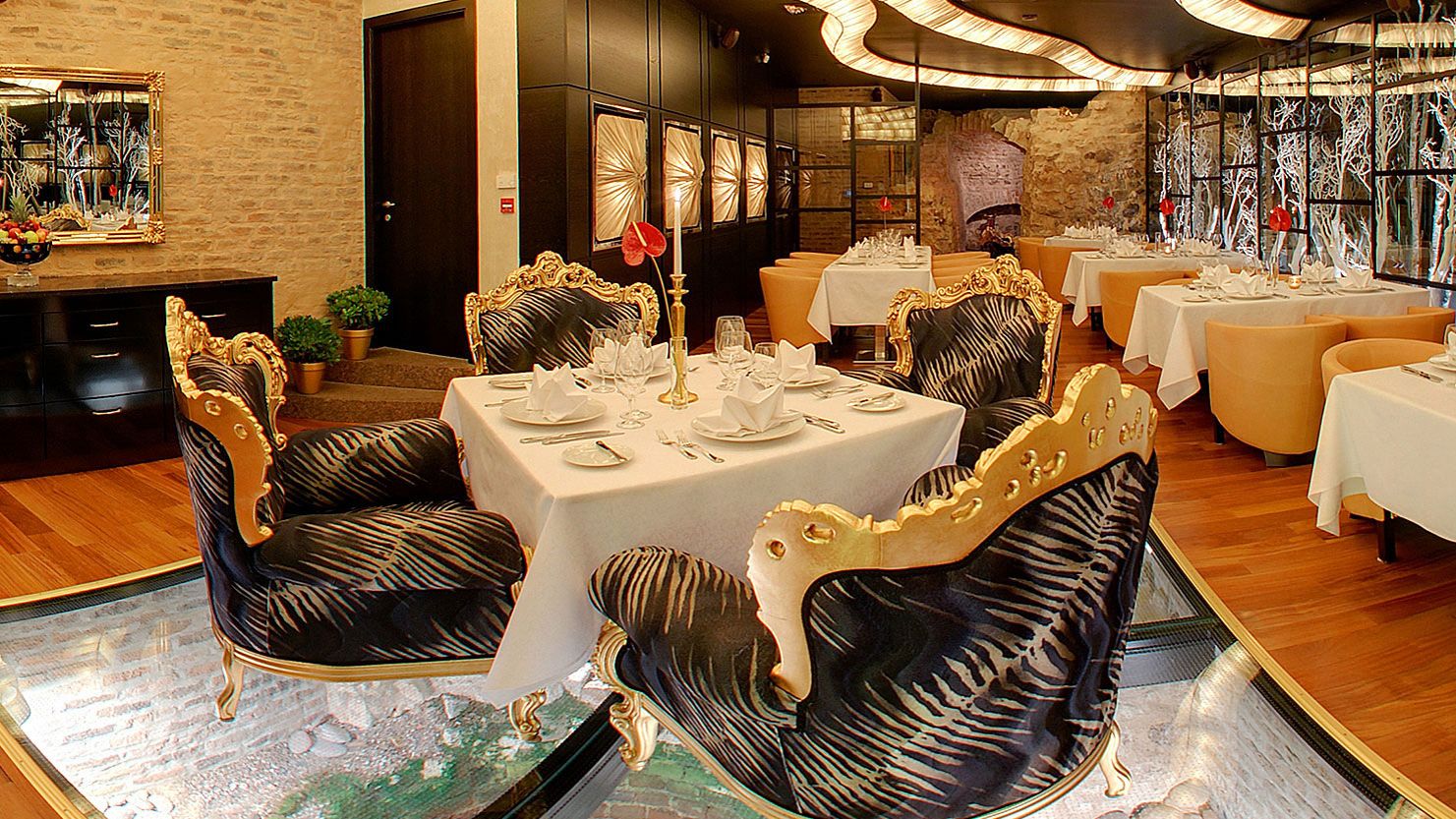 Un restaurante con sillas ornamentadas en negro y dorado, manteles blancos y un fondo de pared de piedra.