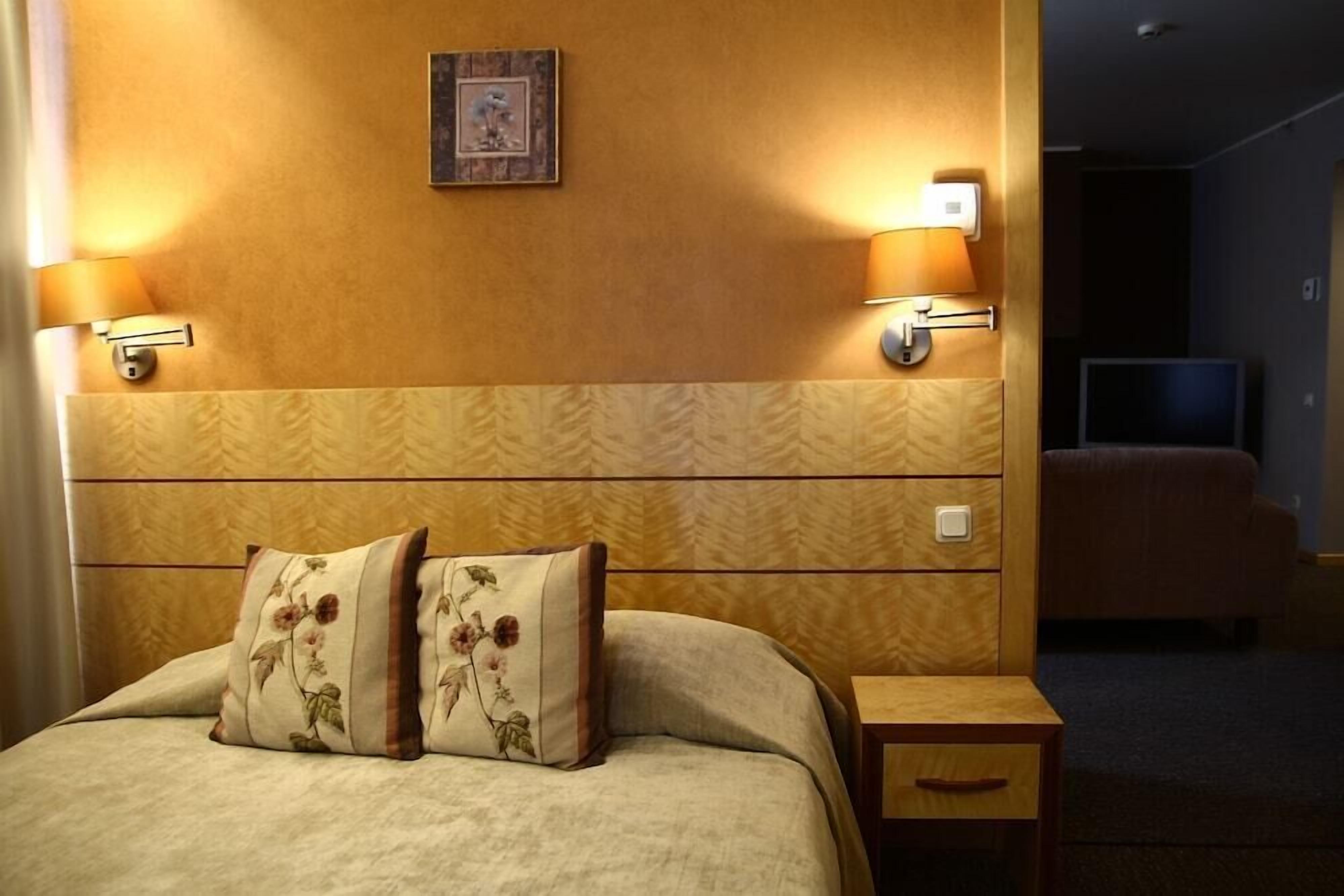 A hotel room with a bed, two wall-mounted lamps, a small bedside table, and a television in the background.