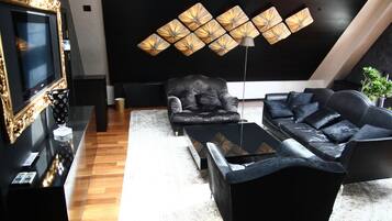 Una sala de estar moderna con un sofá de cuero negro, un sillón a juego y una mesa de centro. A la izquierda, un cuadro grande enmarcado y una lámpara de pared sobre el sofá.