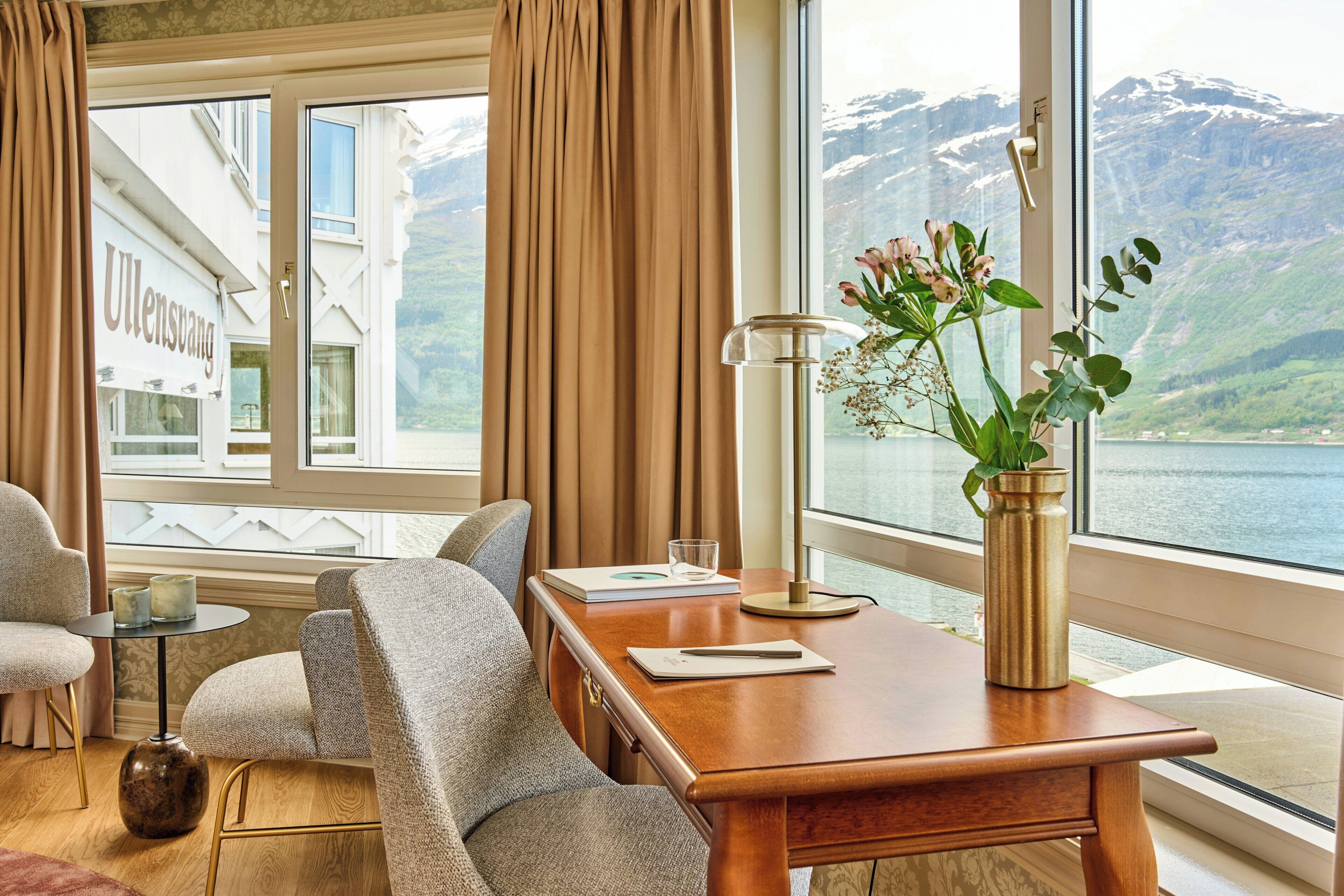 A room with a wooden desk, two chairs, a vase with flowers, a lamp, and a view of a mountain and lake.