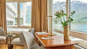 A room with a wooden desk, two chairs, a vase with flowers, a lamp, and a view of a mountain and lake.