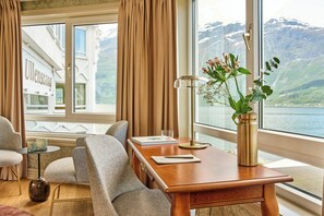 A room with a wooden desk, two chairs, a vase with flowers, a lamp, and a view of a mountain and lake. - Hotel Ullensvang (906914) (Lofthus)