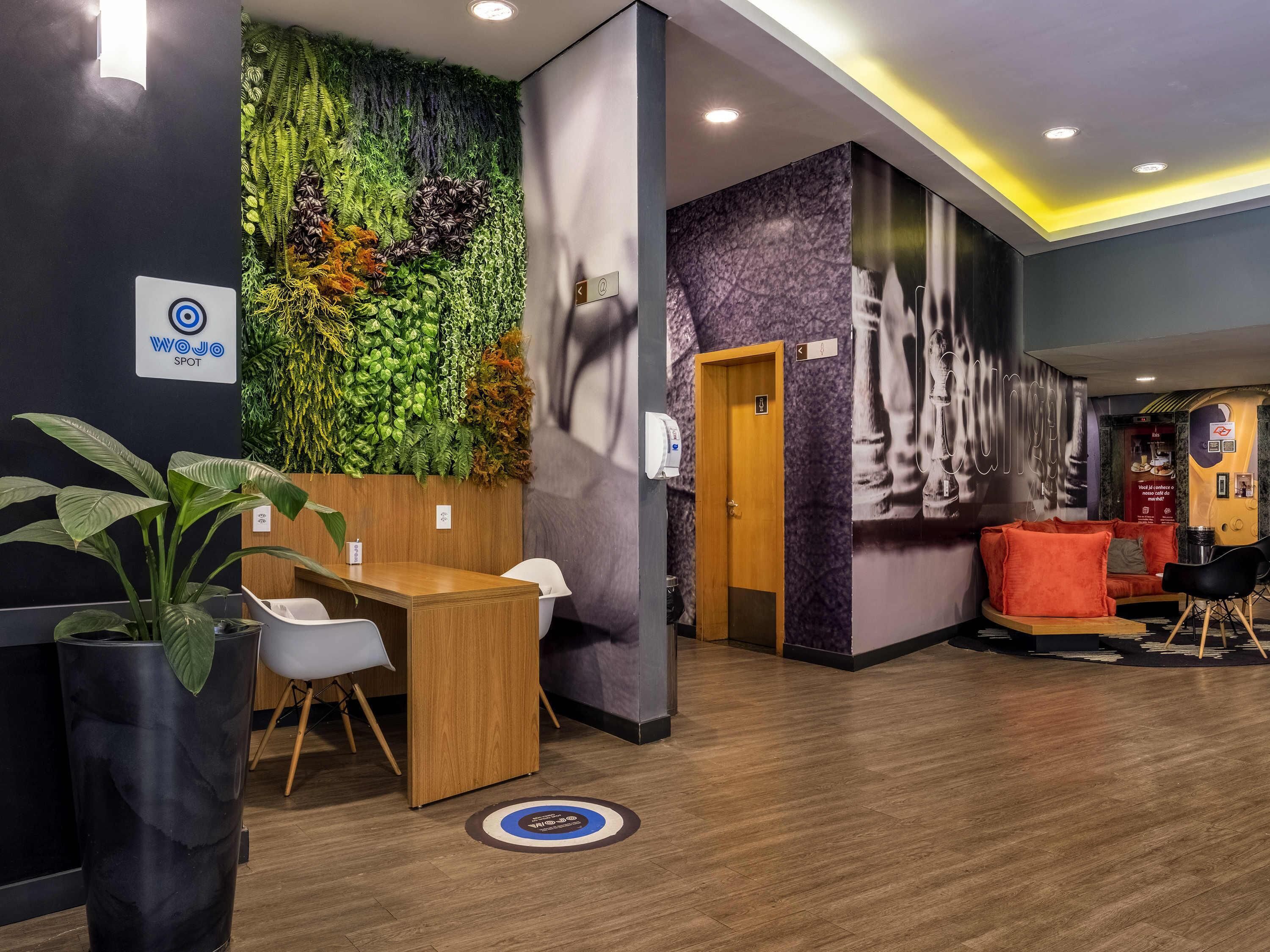 Un espace d’accueil avec un bureau en bois, des chaises blanches, un mur vert orné de plantes et un coin salon avec des sièges orange.