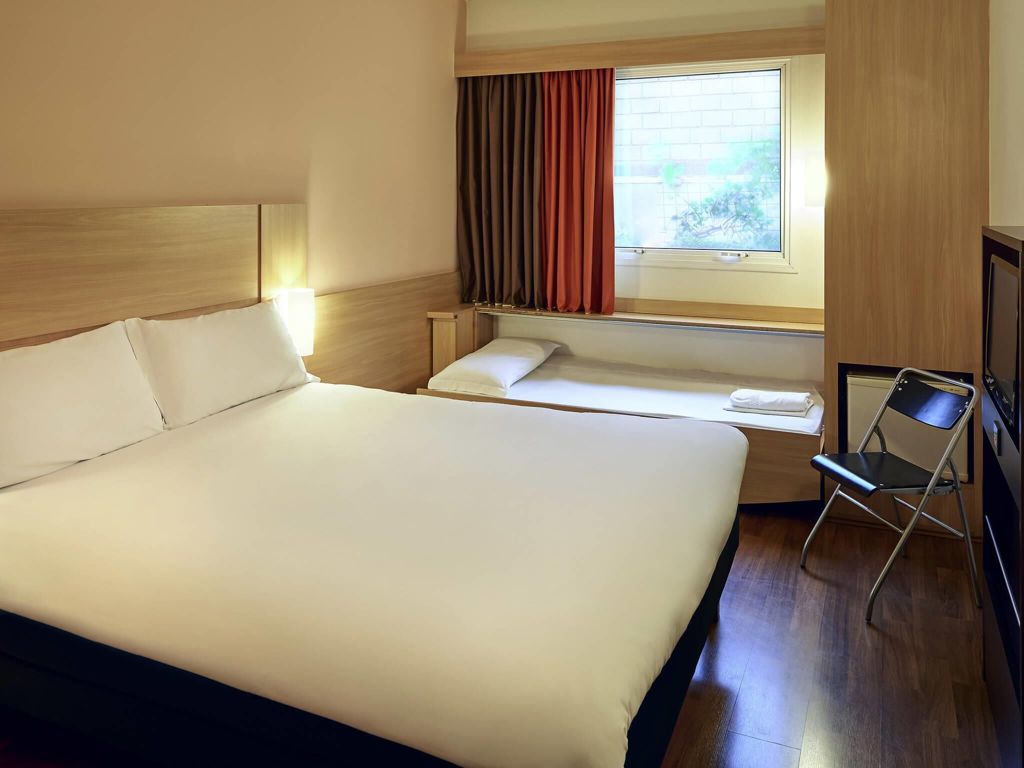 Quarto de hotel com uma cama grande, uma cama menor, uma mesa com cadeira, uma janela com cortinas e uma televisão.