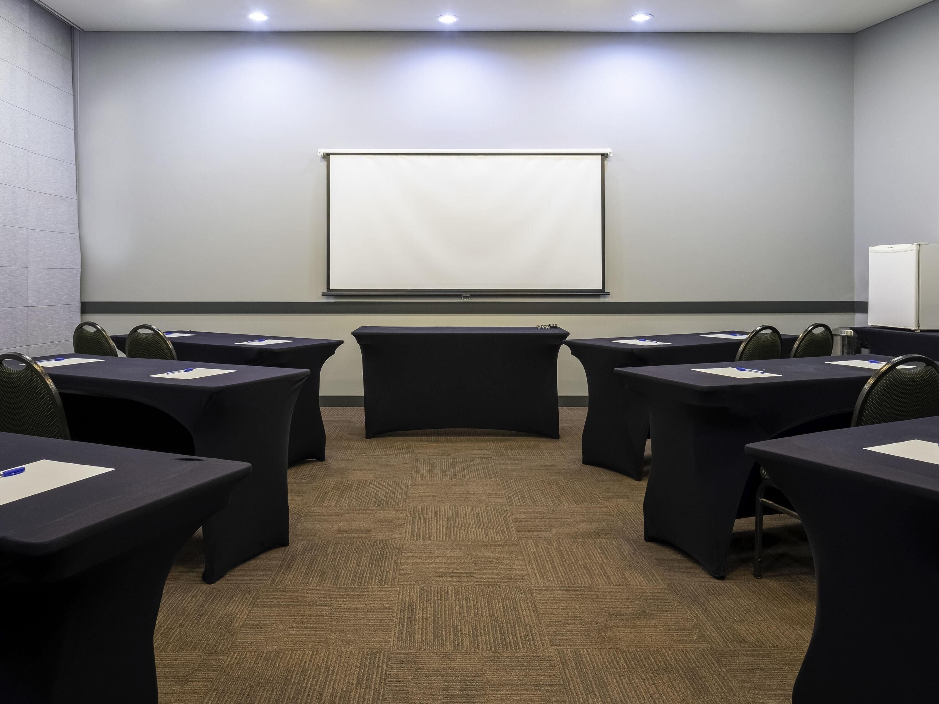 A conference room with a projector screen, tables, chairs, and a refrigerator.