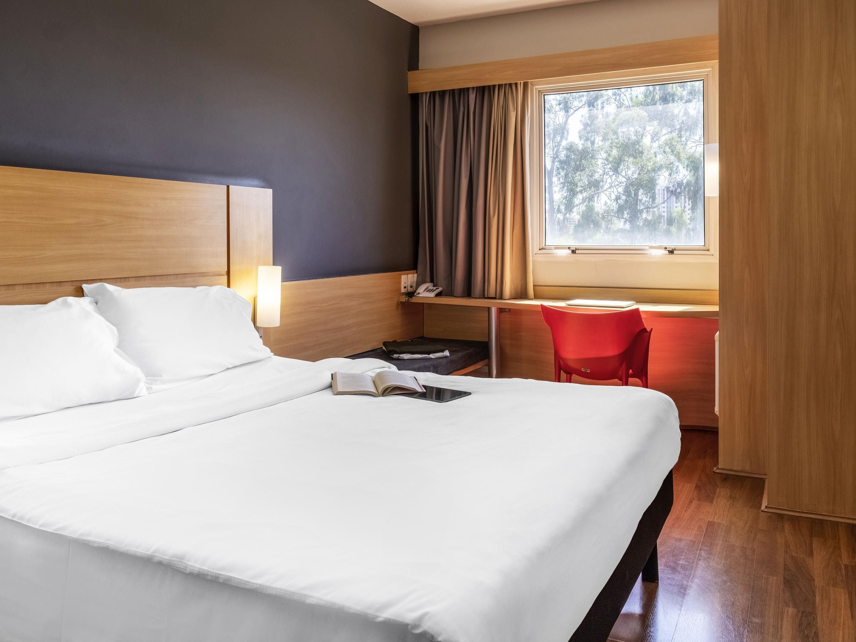 Een hotelkamer met een groot bed, een bureau met een stoel, een raam met gordijnen en een bedlampje.