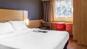A hotel room with a large bed, a desk with a chair, a window with curtains, and a bedside lamp.