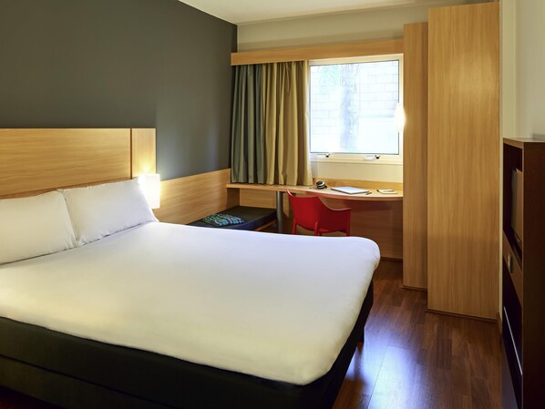 A hotel room with a large bed, a desk with a chair, a window with curtains, and a wardrobe.