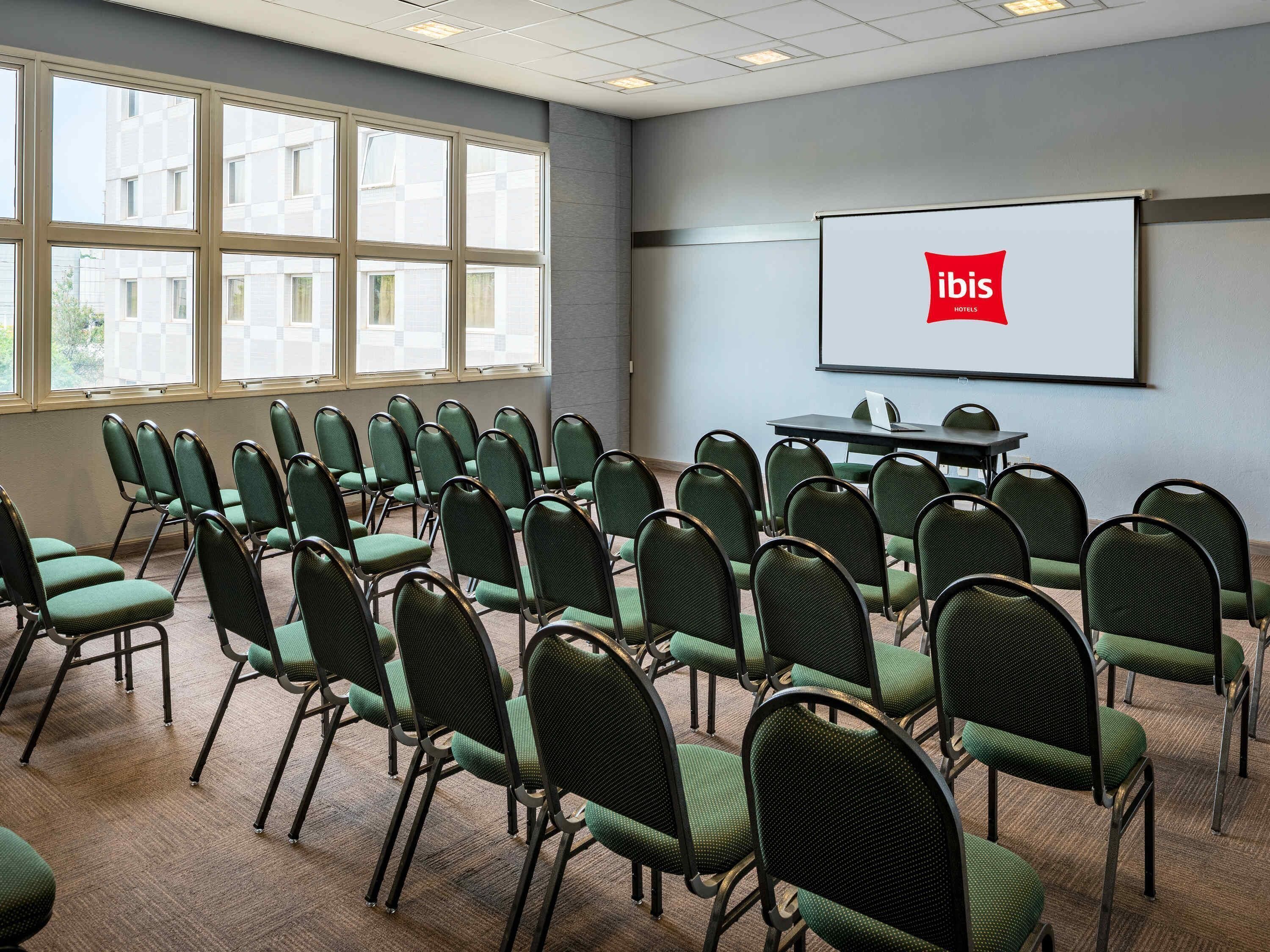 A conference room with rows of green chairs, a projector screen with 'ibis Hotels' logo, and large windows.