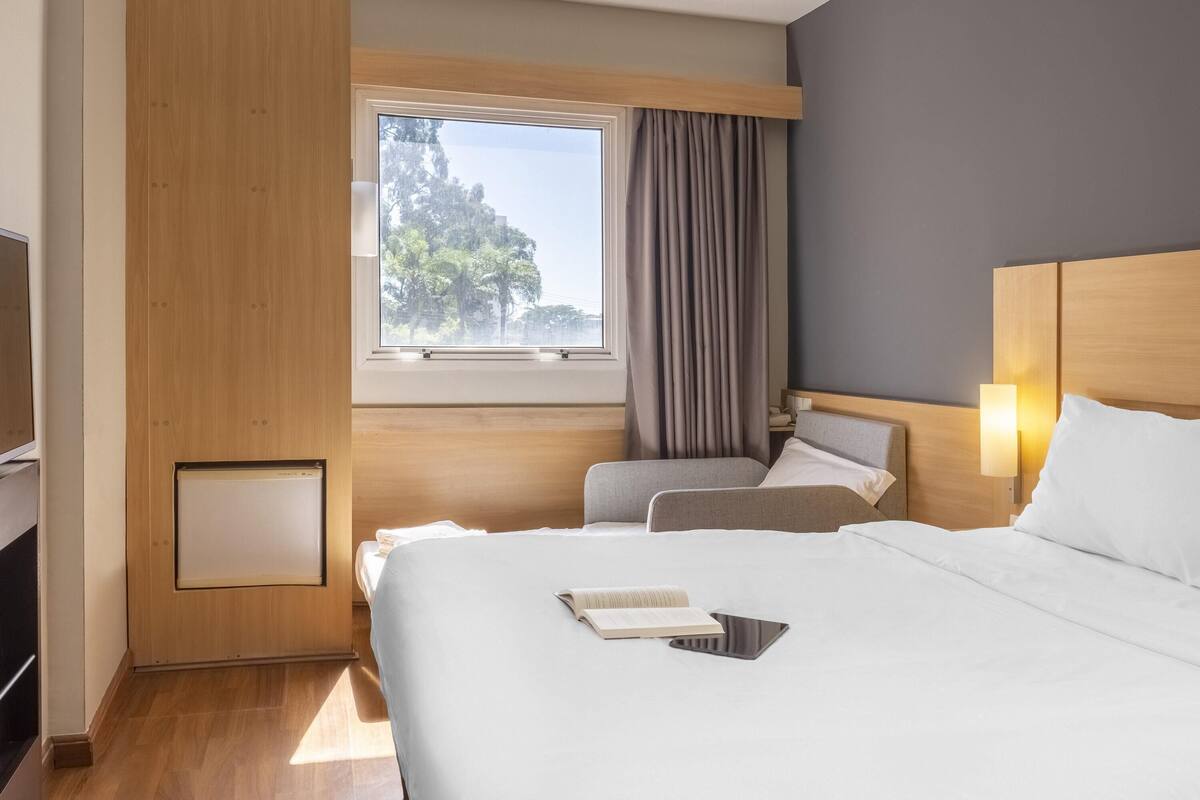 Ett hotellrum med en stor säng, en mindre säng, ett skrivbord, en stol, ett fönster med gardiner och en vägglampa.