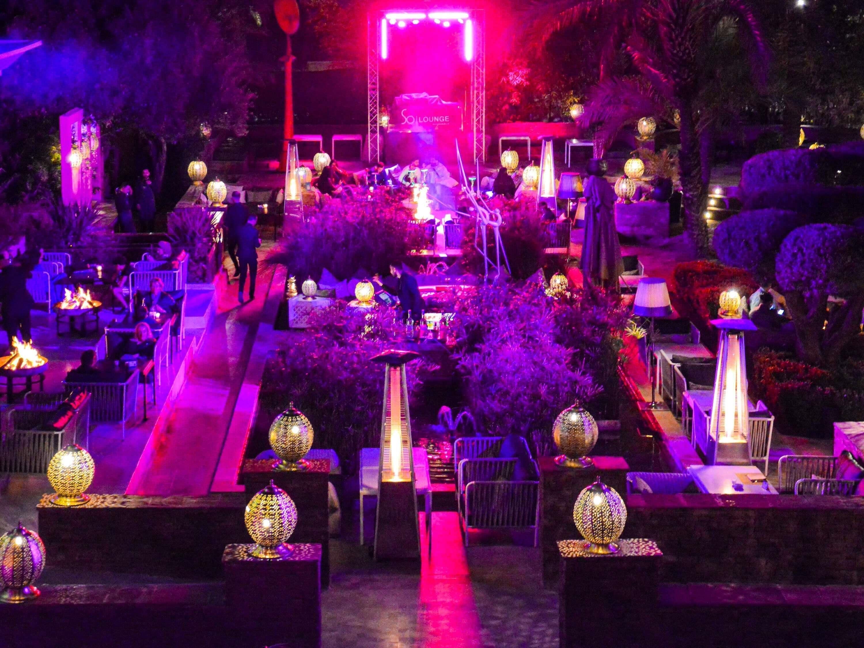 An outdoor dining area with tables and chairs, decorative lanterns, and a stage with a sign.