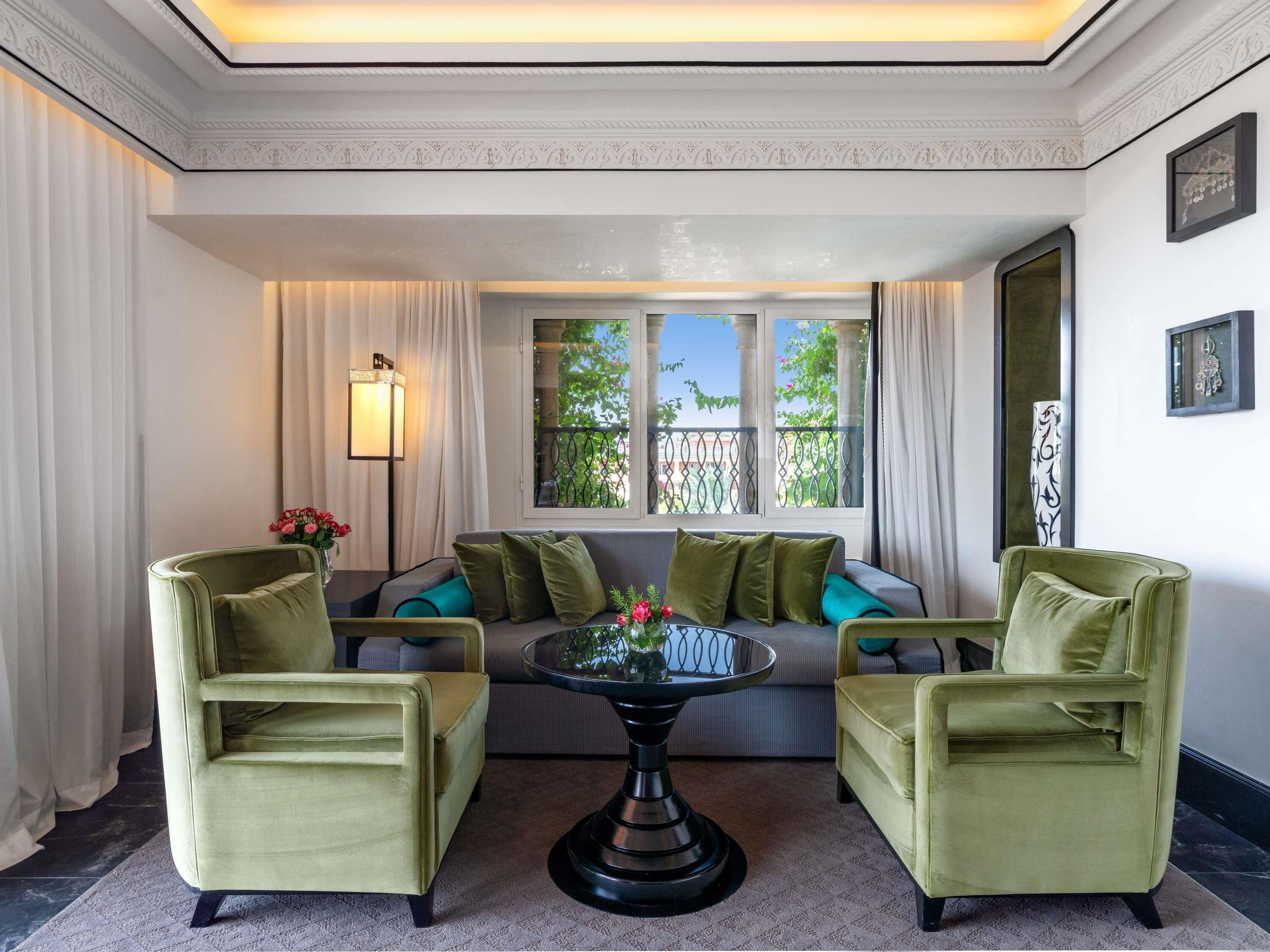 Una habitación con sofá, dos sillones, una mesa con flores, una lámpara y una ventana con vistas a zonas verdes.