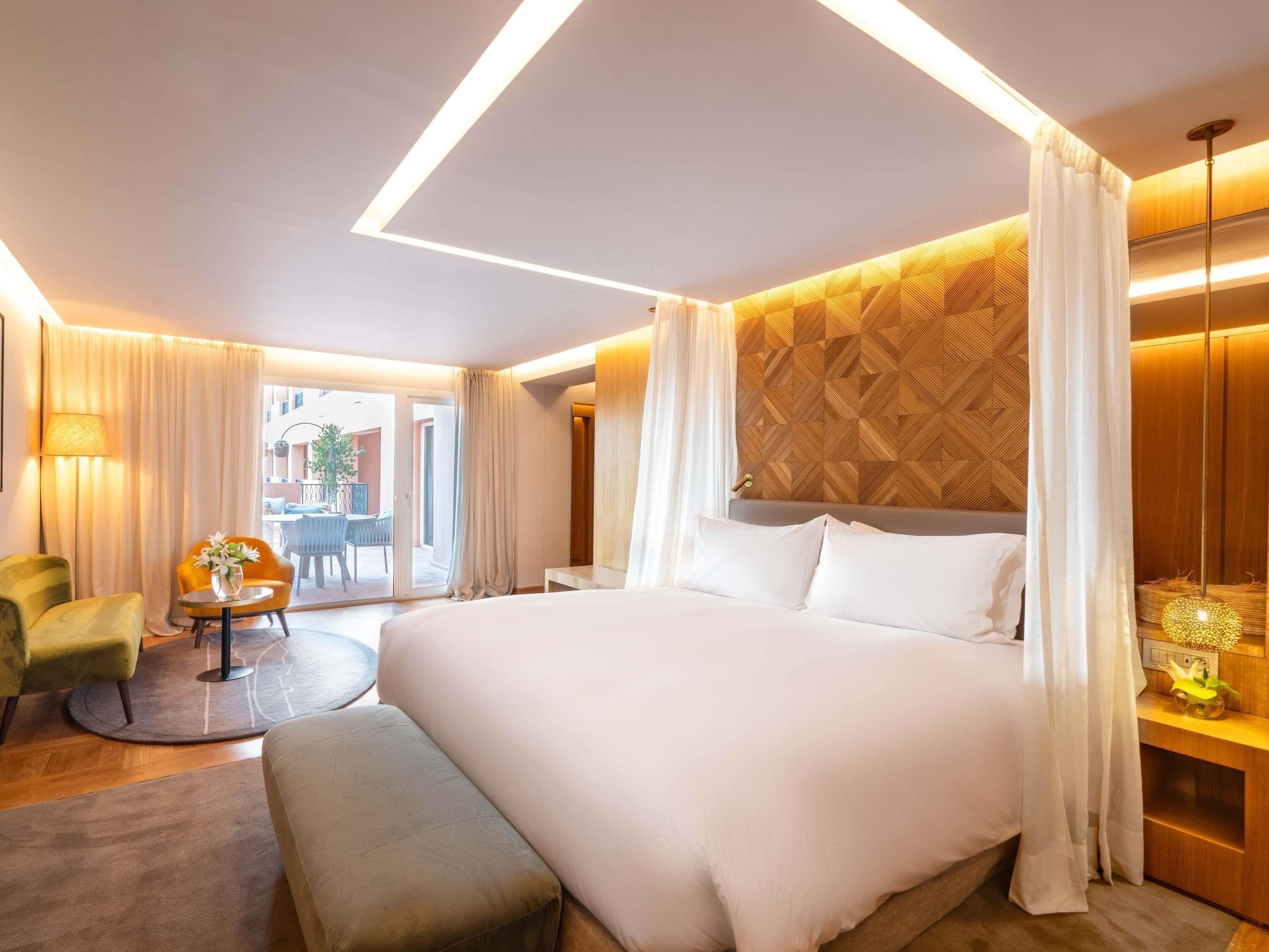 Habitación de hotel moderna con una cama grande, una mesita redonda, un sillón verde y vistas al exterior.