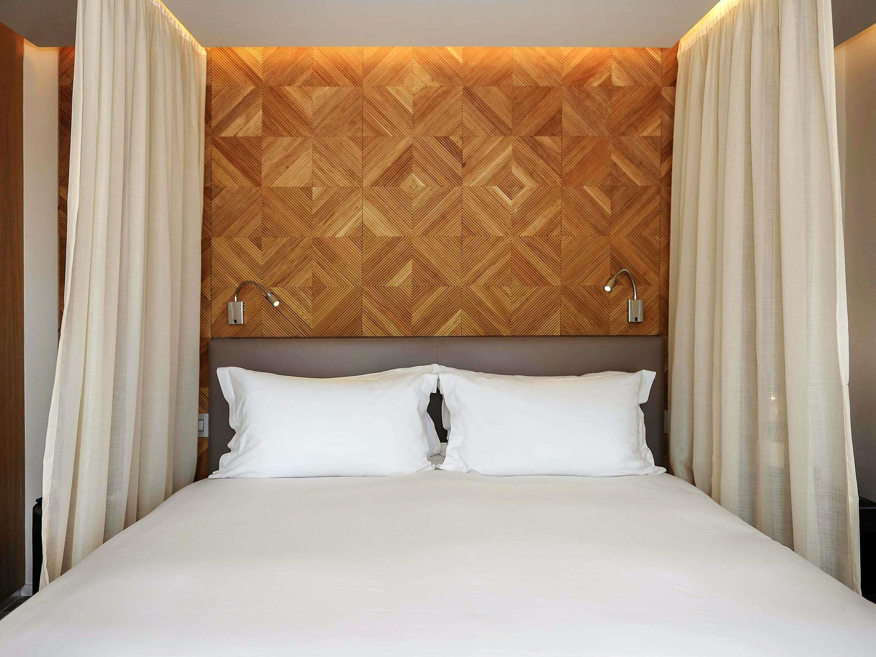 A hotel room with a bed, white curtains, a geometric patterned headboard, and two bedside lamps.