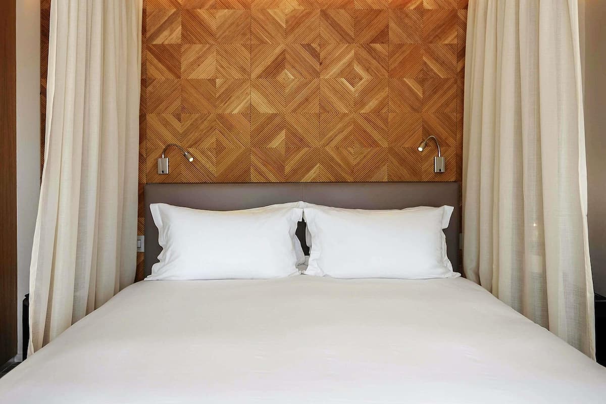 A hotel room with a bed, white curtains, a geometric patterned headboard, and two bedside lamps.