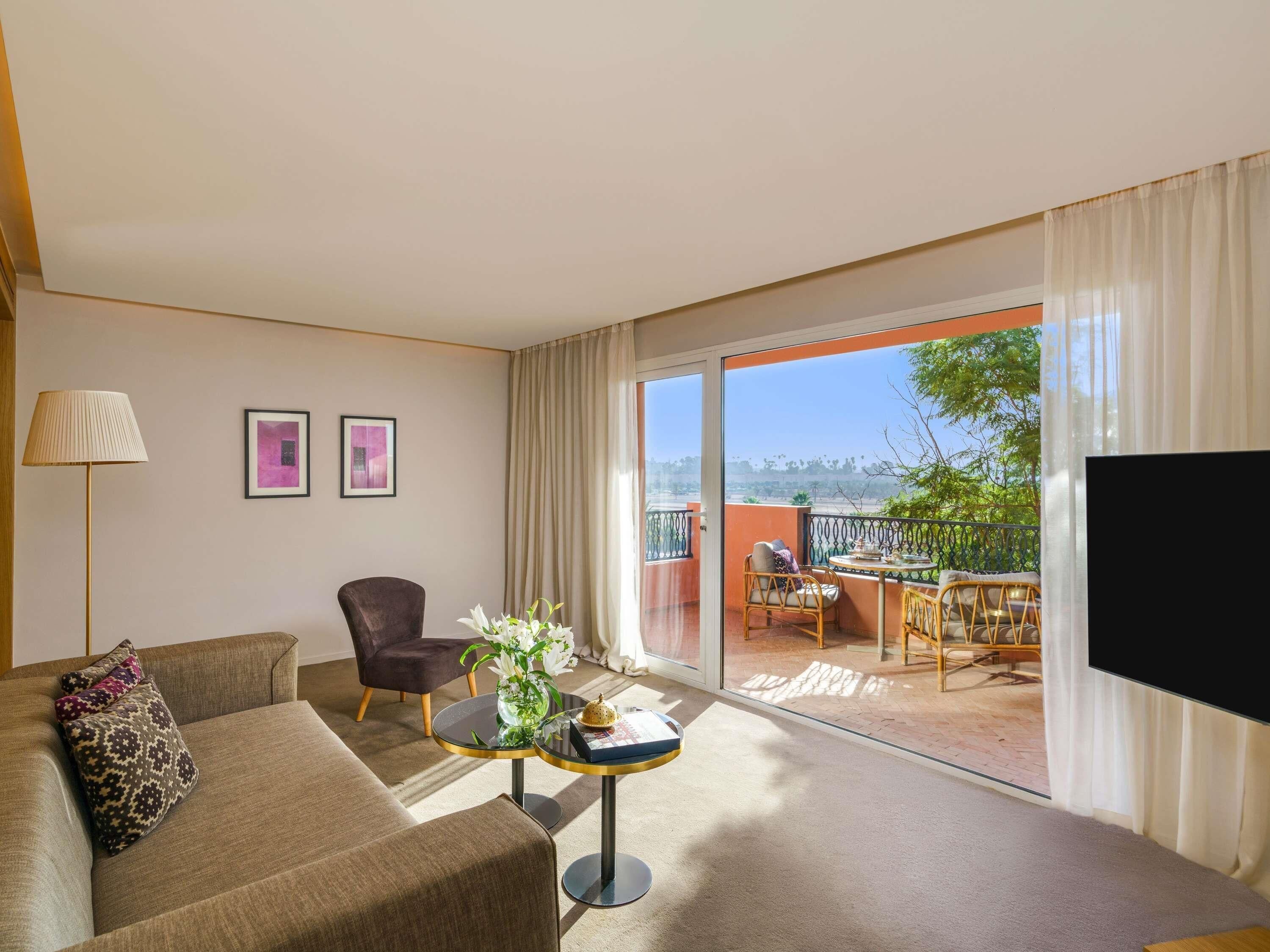 Habitación de hotel con sofá, sillón, una mesita con un jarrón y balcón con mobiliario exterior.