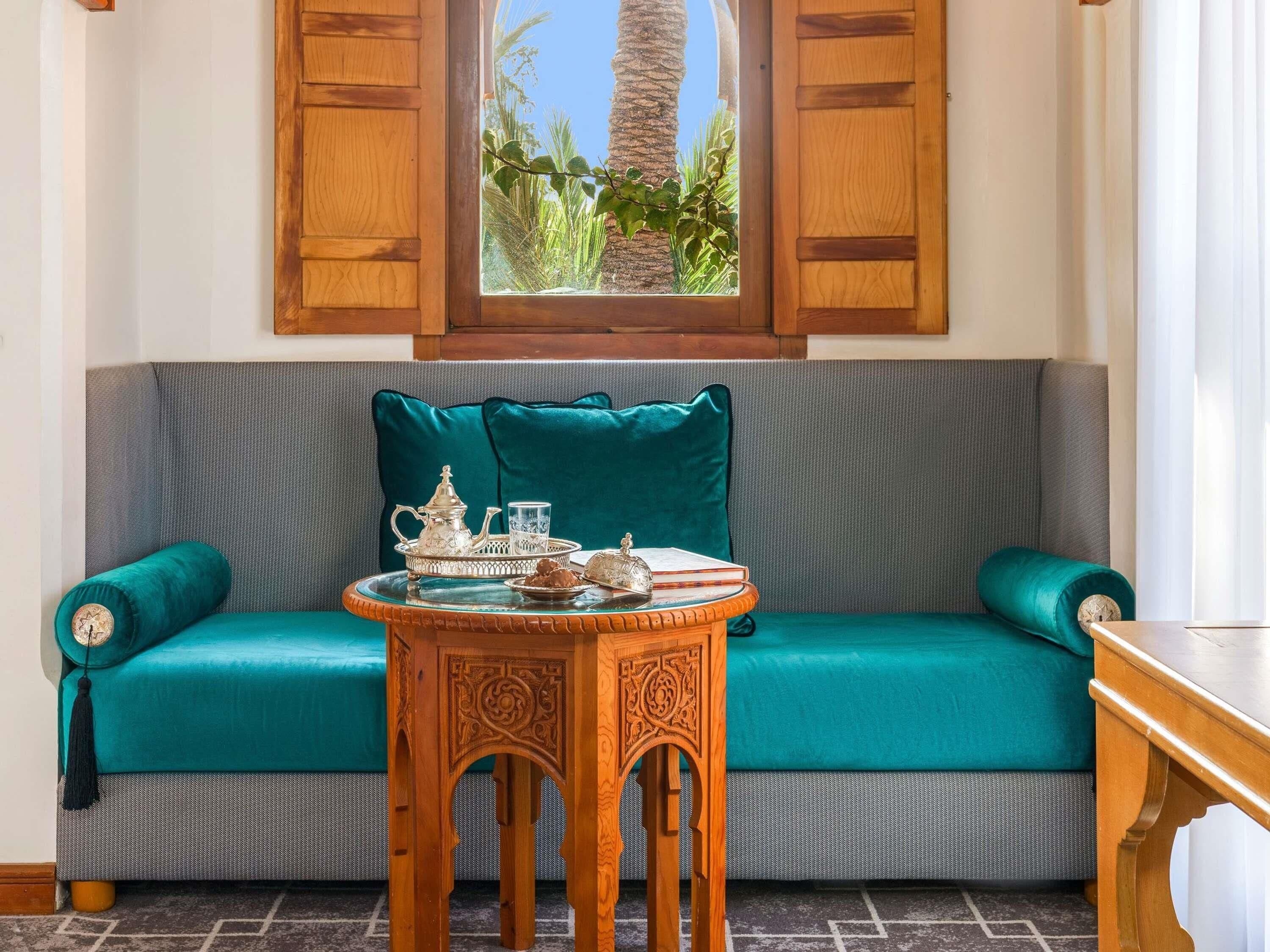 Un sofá color turquesa con vistas a palmeras a través de una ventana, una mesita de madera con una bandeja y un cojín decorativo.