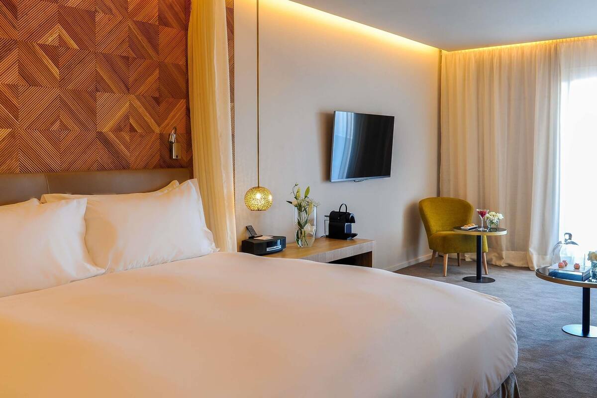 A hotel room with a large bed, a flat-screen TV, a small table with a chair, and a decorative wall panel.