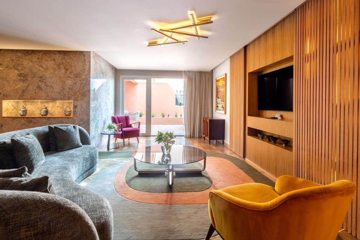 A modern living room with a sofa, a yellow armchair, a glass coffee table, a flat-screen TV mounted on a wooden panel, and a view of the outdoor balcony.