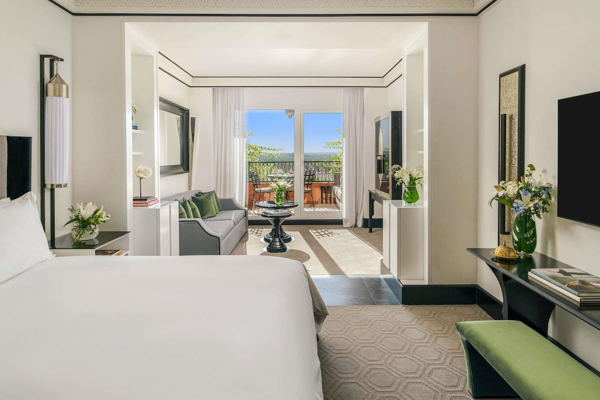 A hotel room with a large bed, a sofa, a TV, a balcony with a view, and a dining area.