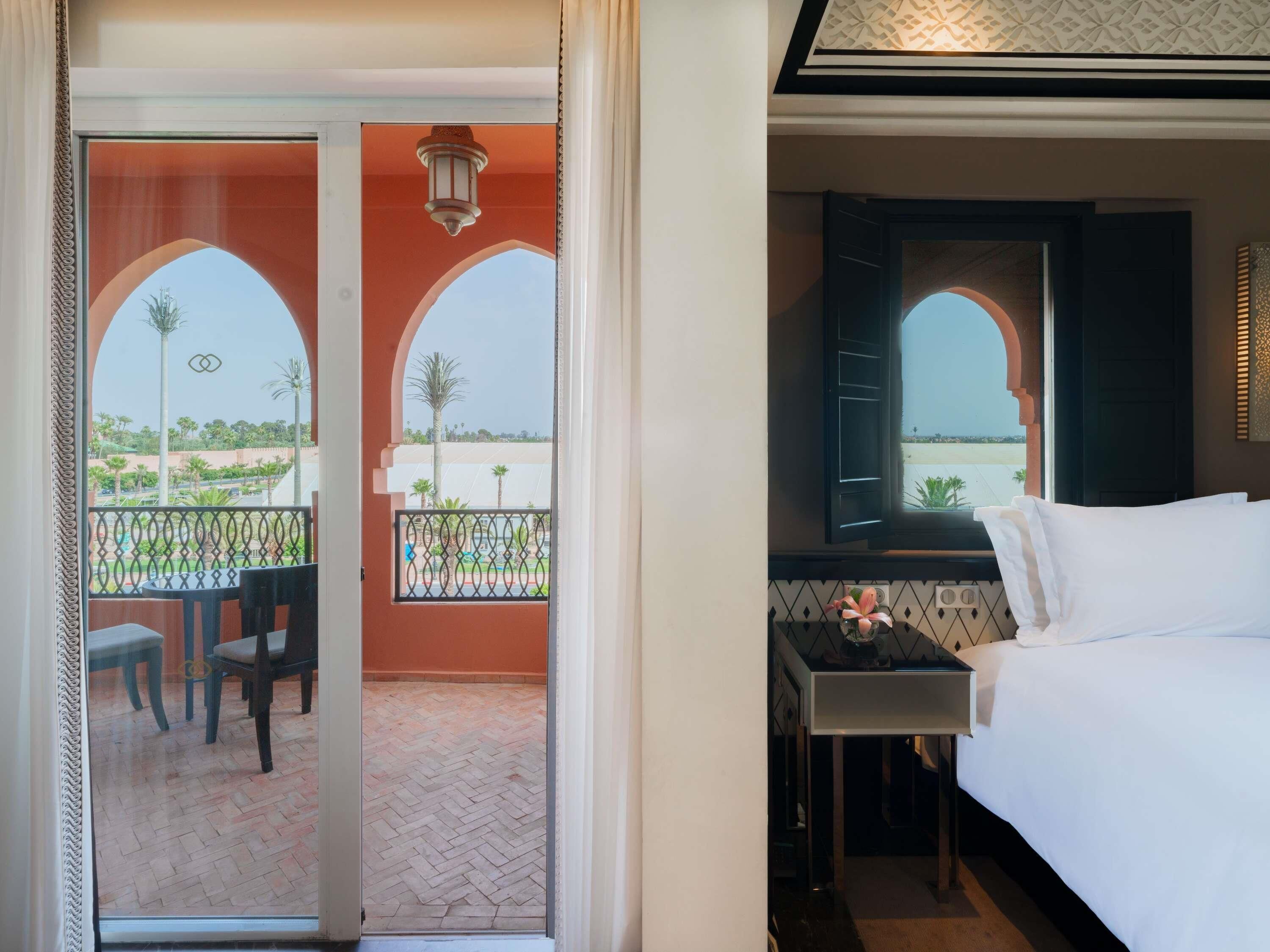 A hotel room with a bed, a small table with a vase, a window with open shutters, and a balcony with a table and chairs.