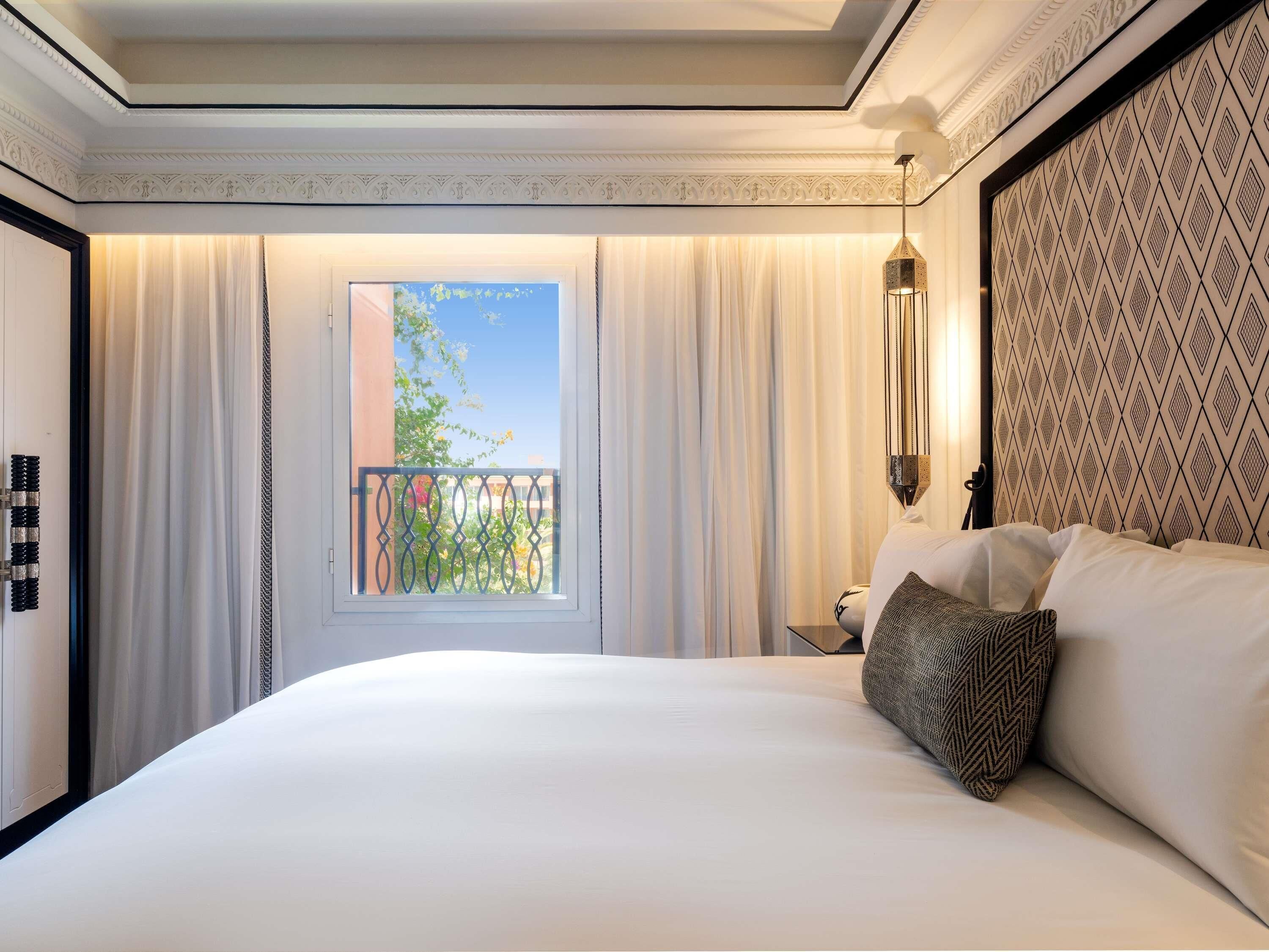 A hotel room with a large bed, a window with curtains, a balcony, and a patterned wall.