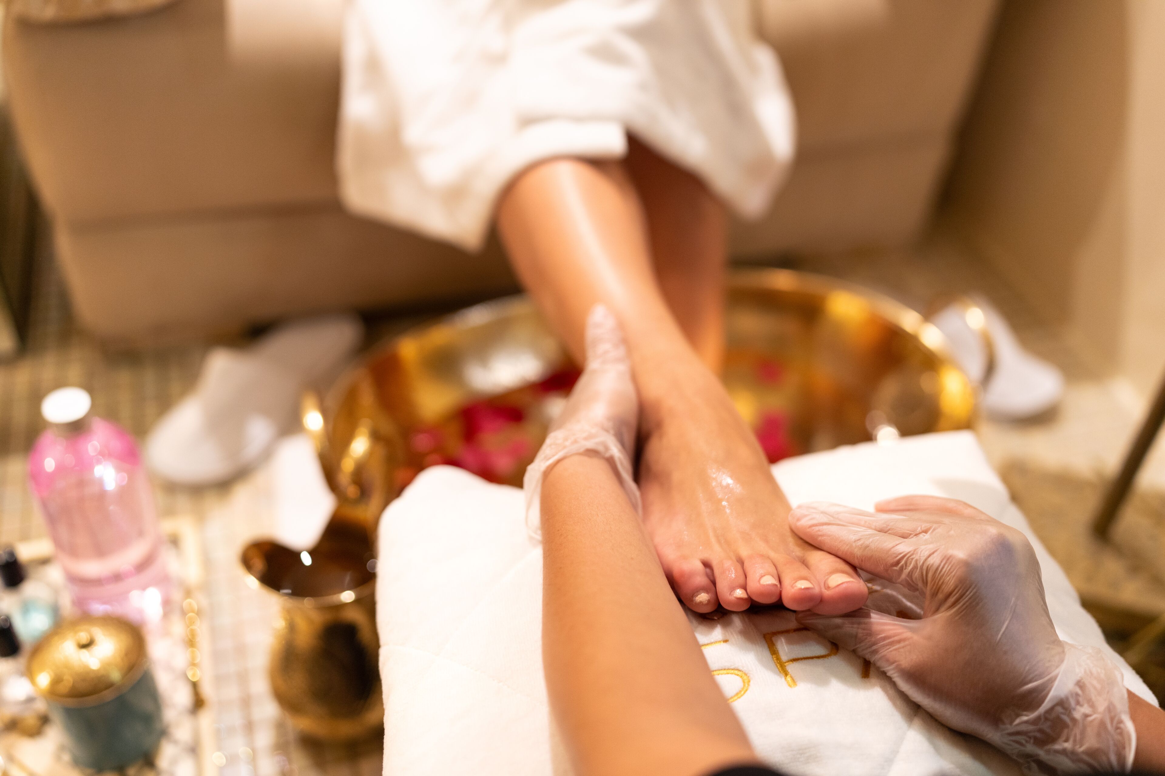 Un massage des pieds accompagné d’un bain de pieds, d’une bouteille de lotion et d’un récipient doré, le tout posé sur un sol carrelé.