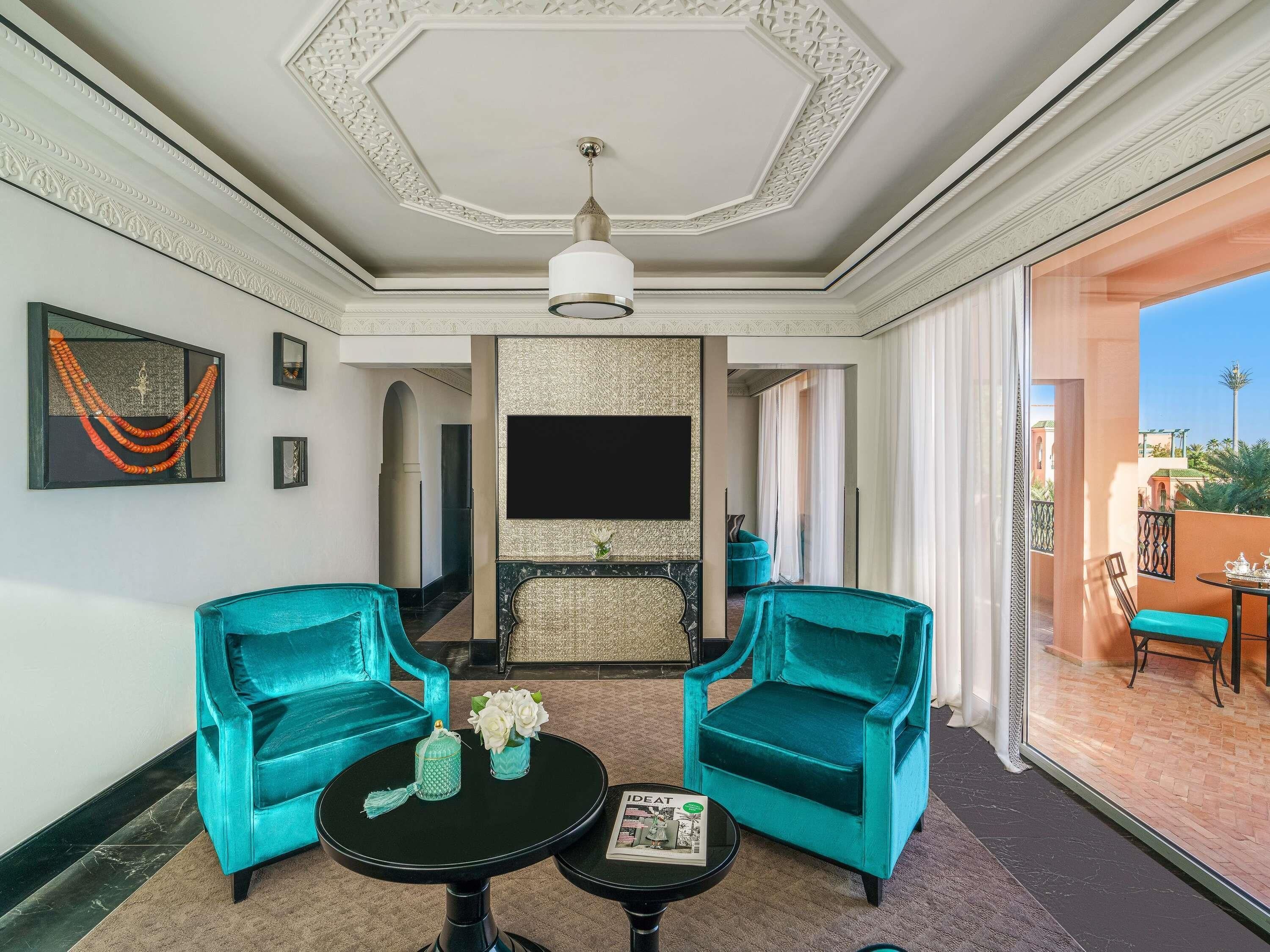 A room with two teal armchairs, a black table, a TV, and a balcony with a table and chairs.