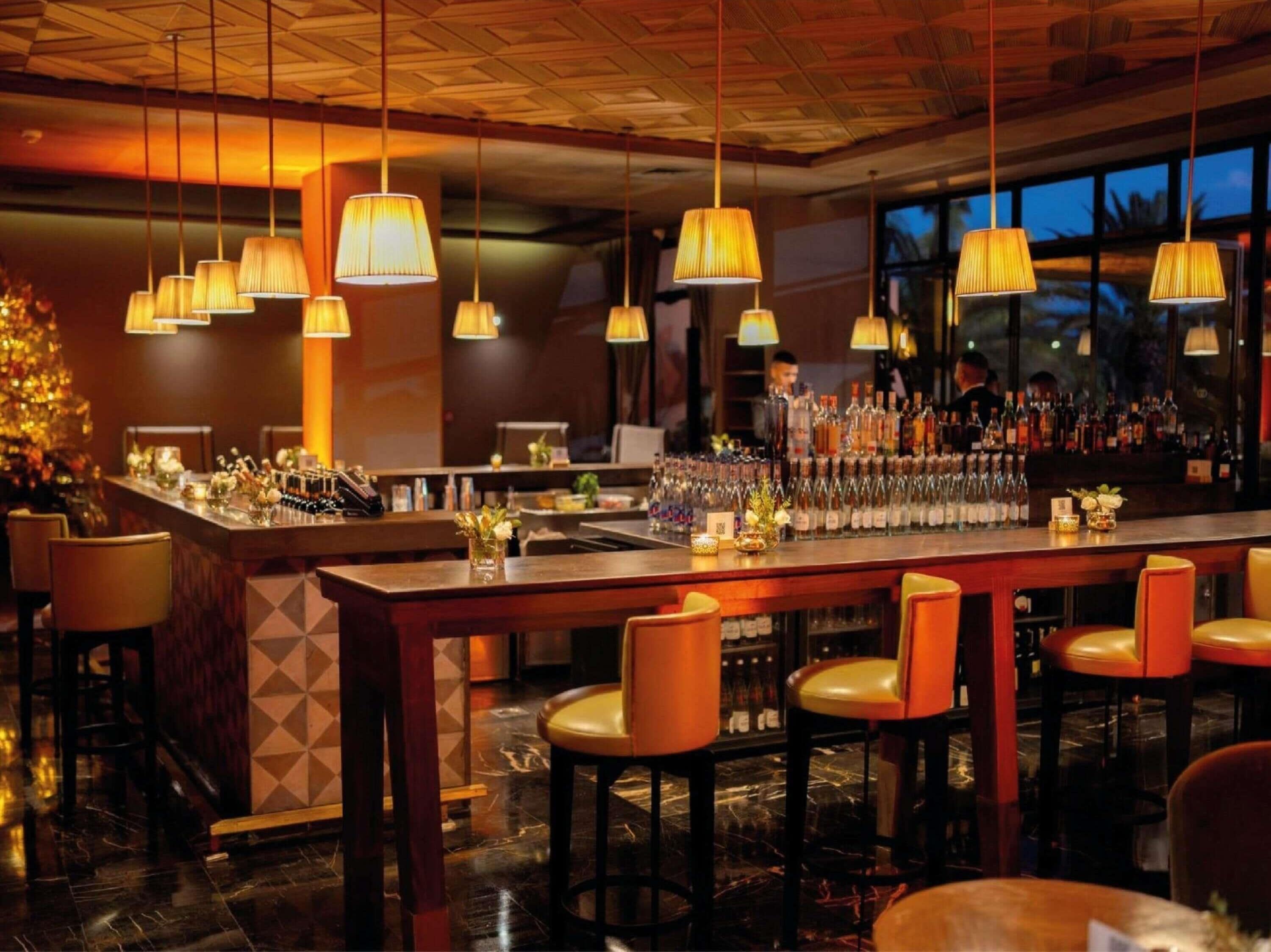 Zona de bar con varias lámparas colgantes, barra con botellas y taburetes.