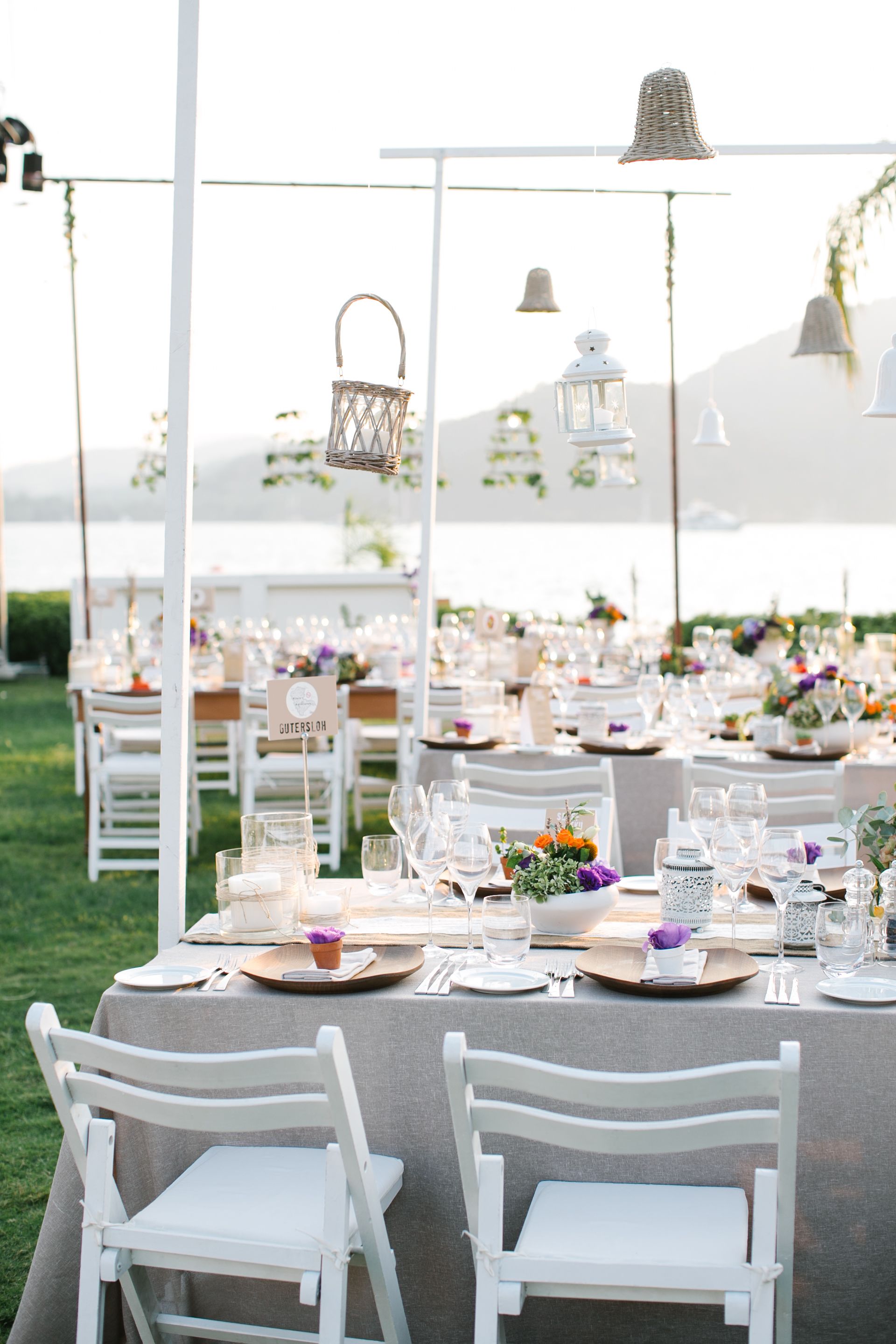 An outdoor wedding reception with white tables and chairs, floral centerpieces, and hanging lanterns.