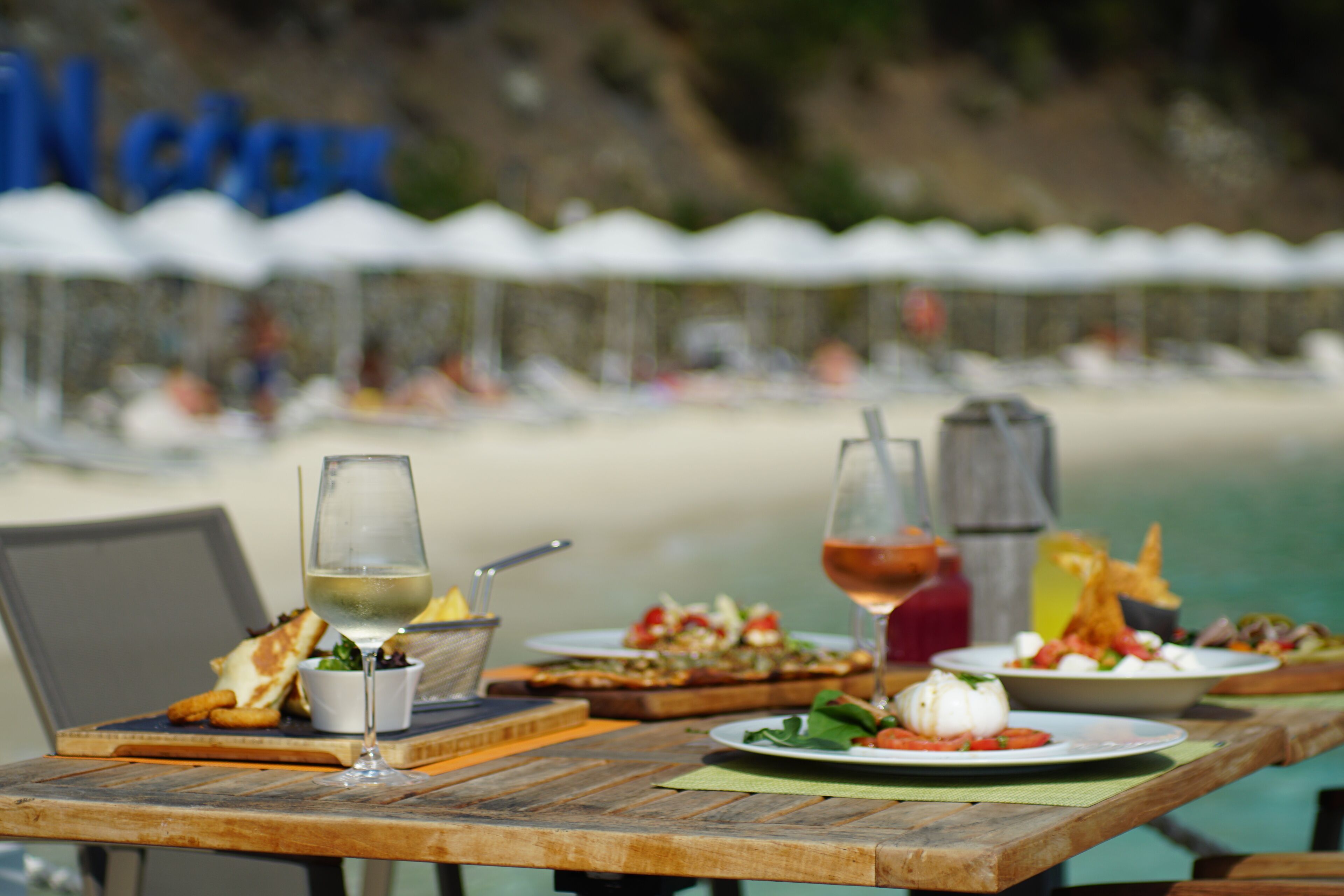 Une table à manger dressée avec différents plats et boissons, le tout installé sur une plage, avec des parasols en arrière-plan.