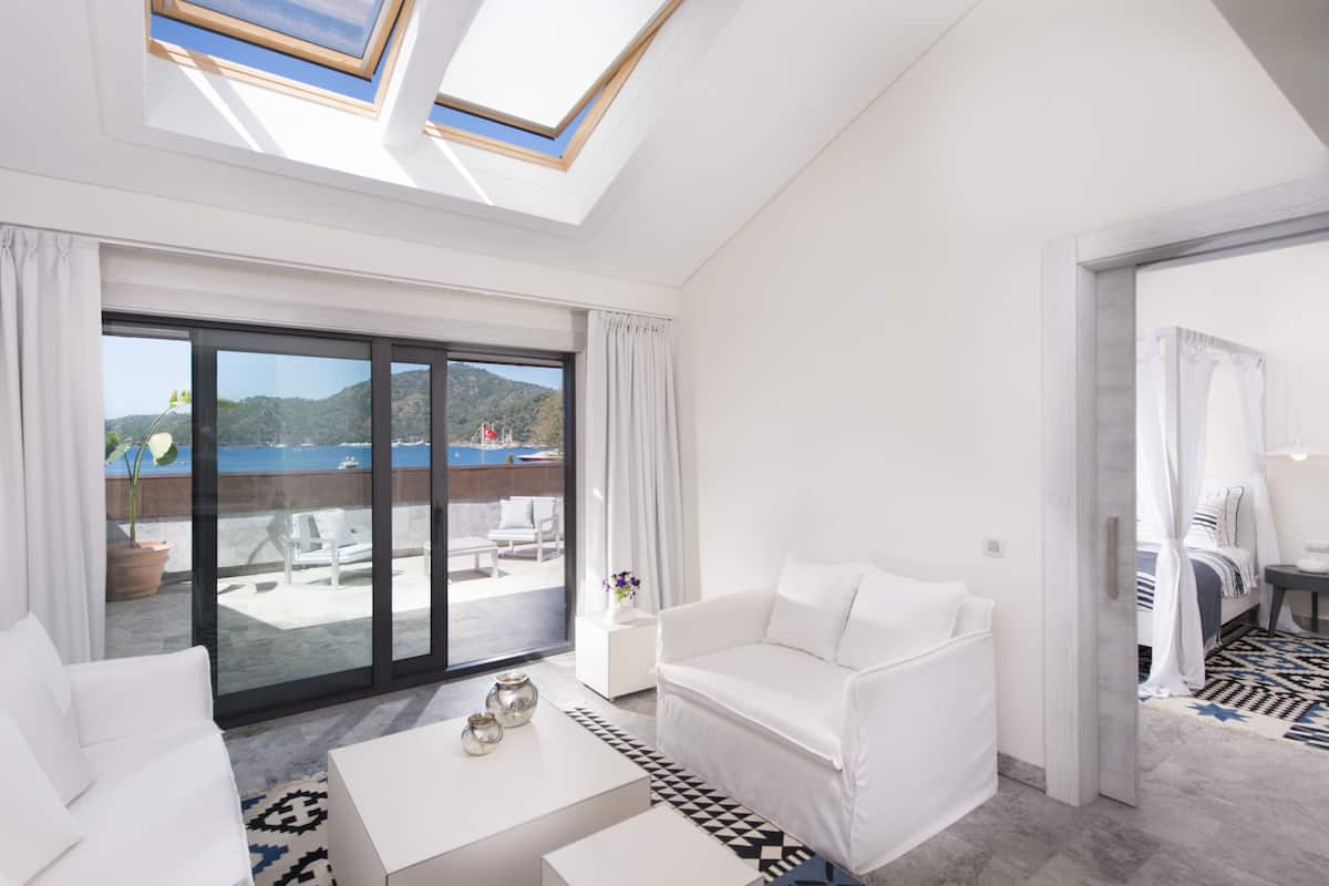 Ein modernes Hotelzimmer mit einer großen Glasschiebetür, die auf einen Balkon mit Blick auf das Meer und die Hügel führt. Es gibt ein weißes Sofa, einen kleinen Couchtisch und ein Himmelbett.