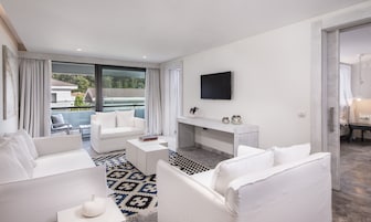 A modern living room with a white sofa set, a coffee table, and a TV mounted on the wall. There is a balcony with a view of the outside.
