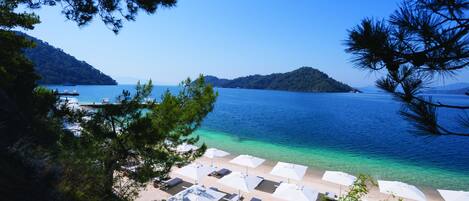 A beach with white umbrellas and lounge chairs, a clear blue sea, and a hilly landscape in the background.