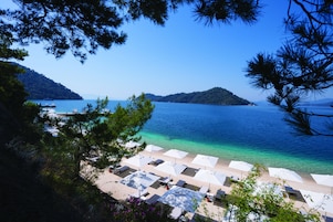 A beach with white umbrellas and lounge chairs, a clear blue sea, and a hilly landscape in the background.