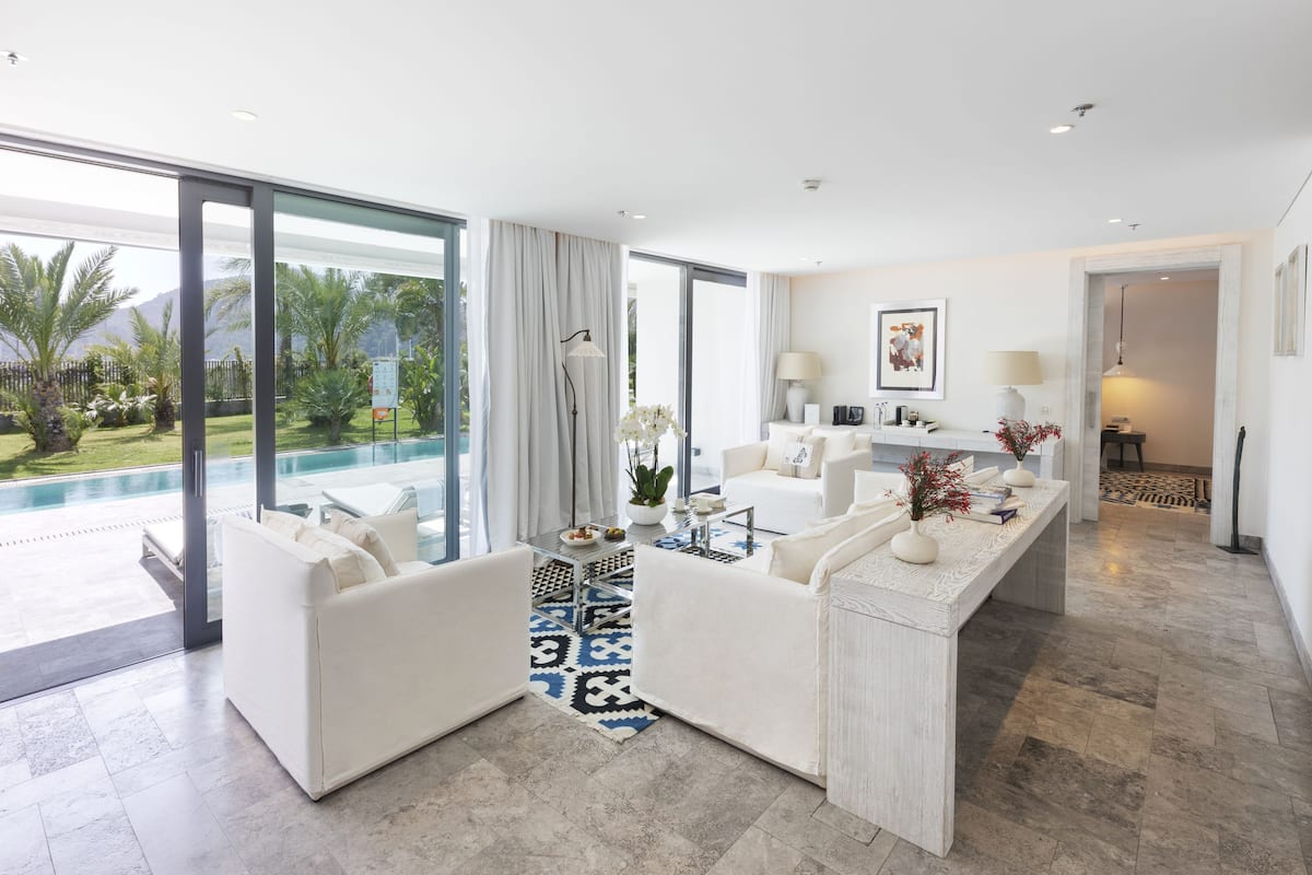 Ein modernes Wohnzimmer mit einem großen Glasfenster zum Pool, einem weißen Sofa, einem Couchtisch und einer kleinen Küchenzeile mit einer Lampe und einem gerahmten Bild an der Wand.