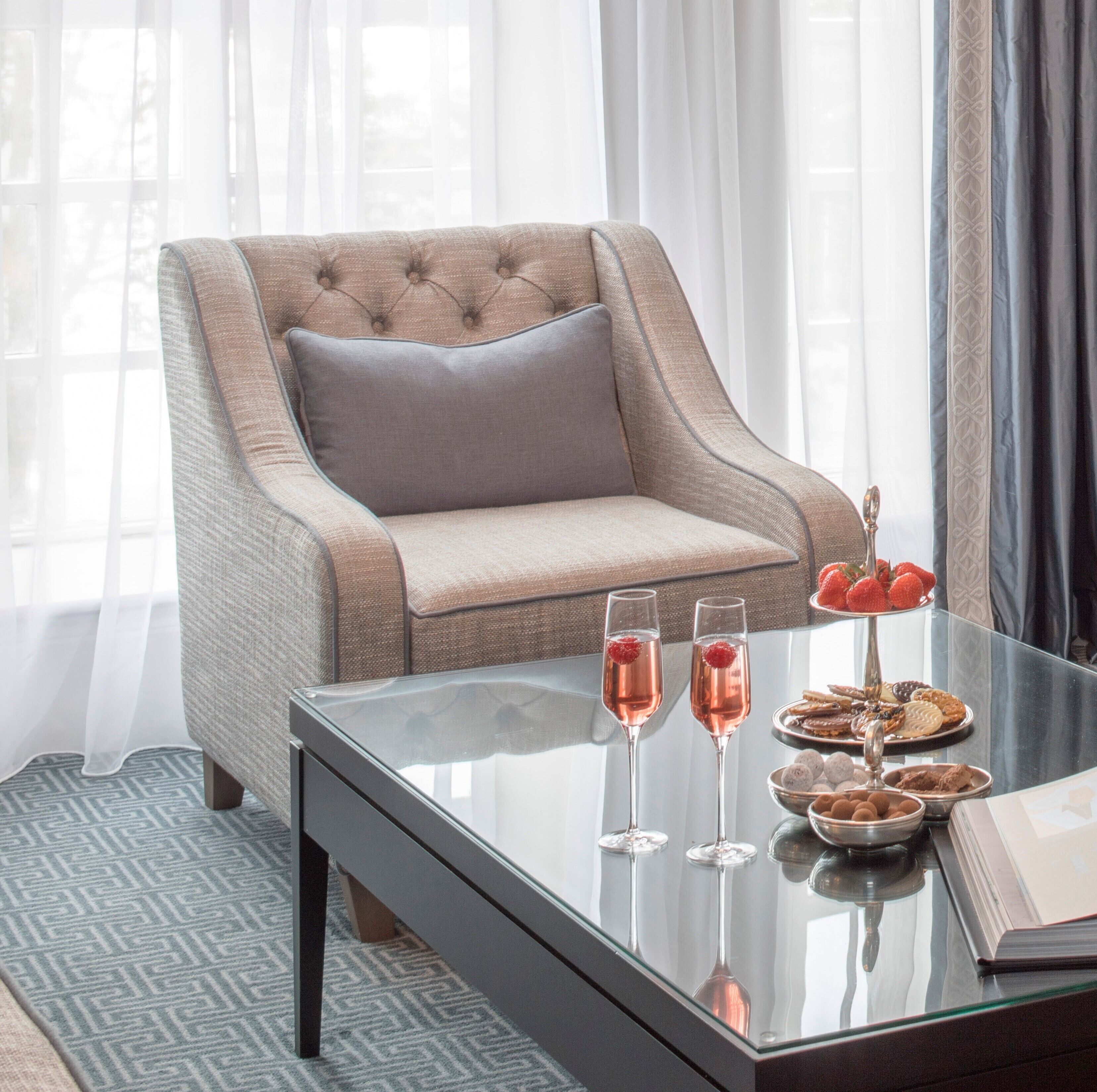A modern living room with a tufted armchair, a glass coffee table with snacks and drinks, and sheer curtains allowing natural light.