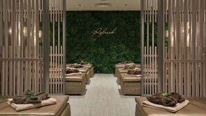 Entrada de spa con una pared verde y la palabra "Refresh" en el centro. Hay sillones de mimbre con toallas y una pequeña planta en cada uno.
